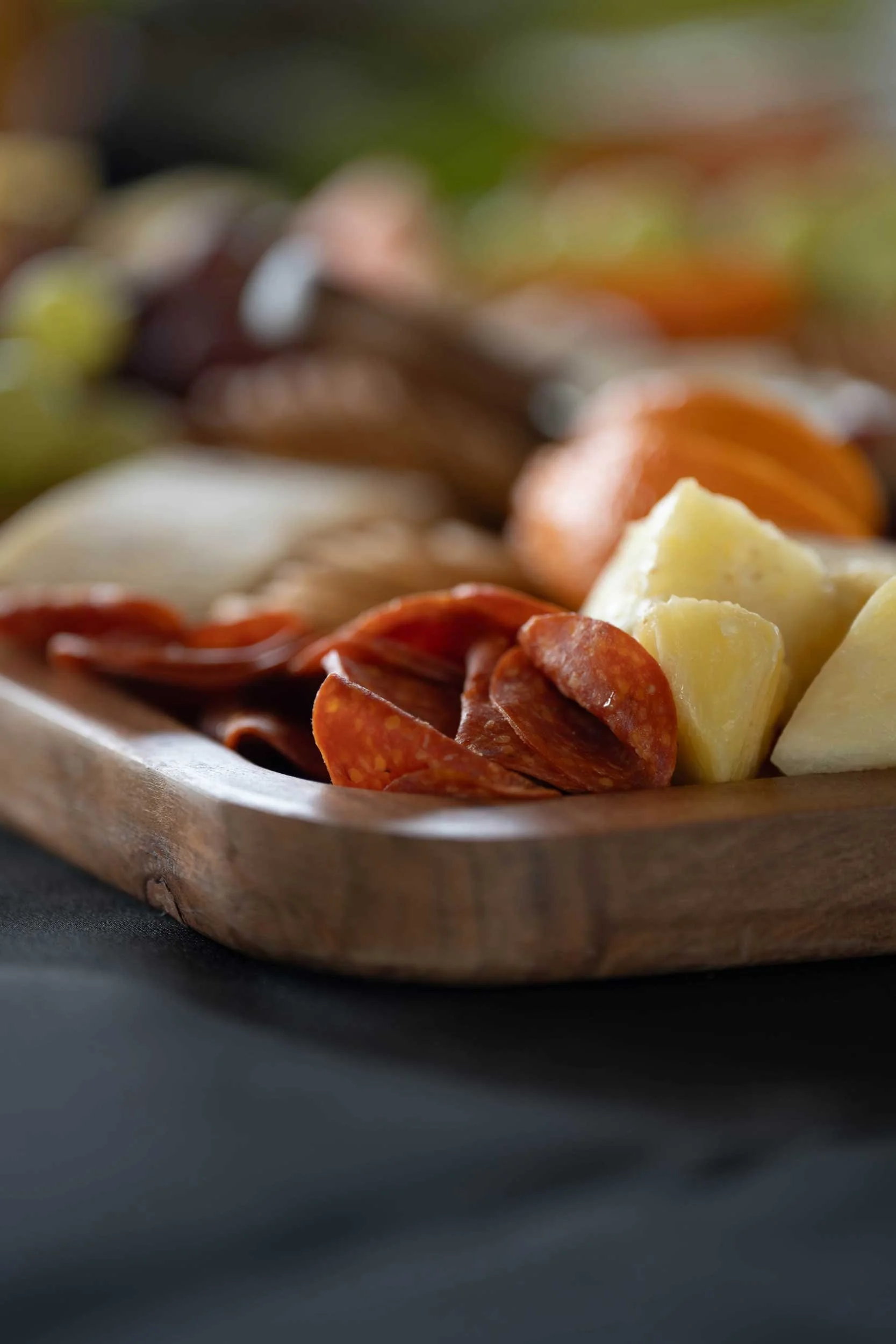 Close-up of a wooden platter with sliced pepperoni, chunks of cheese, and sausages, on a dark background.