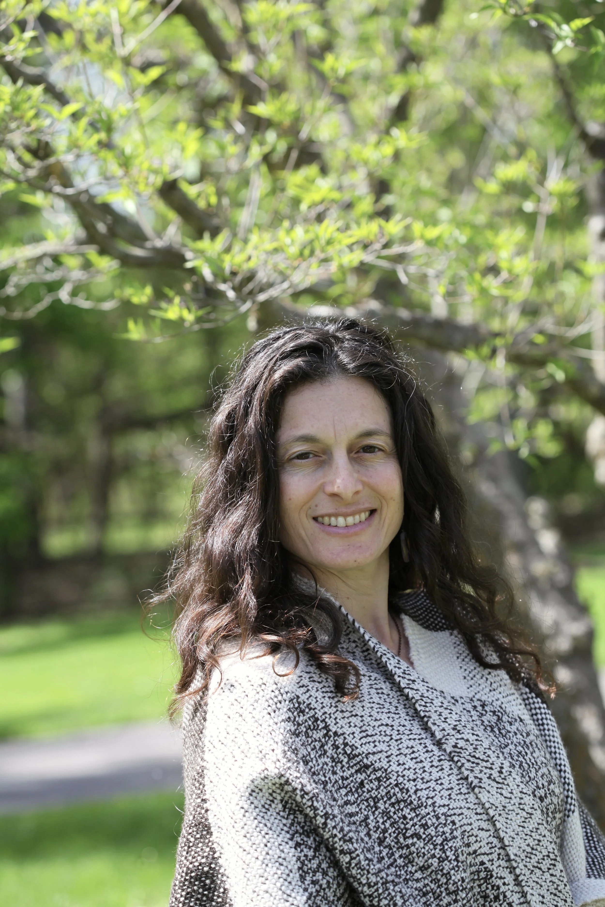 A smiling woman with long curly dark hair stands outdoors in a park with green trees and grass, wearing a patterned black and white sweater.