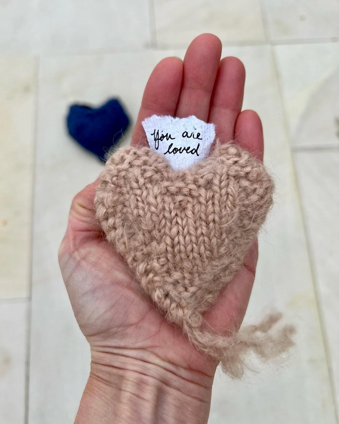 Sara Guren's hand holding a pink hand-knitted heart with a small piece of torn hand-made paper that says 'You are loved' in handwriting, with a blurry blue heart in the background on an outdoor patio.