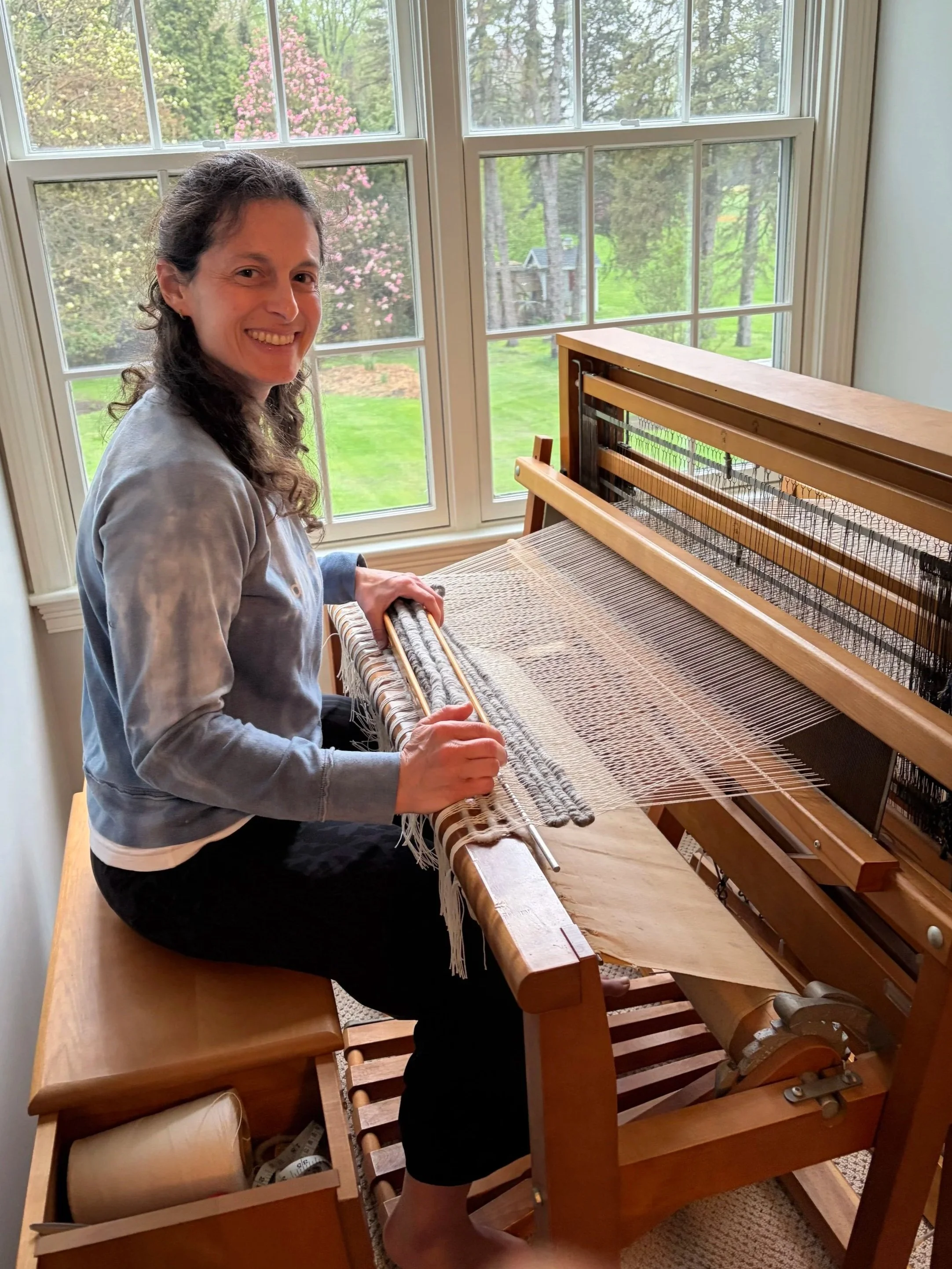 Sara Guren seated at a loom in front of large windows with a view of green trees and pink flowering trees outside, weaving a rug.