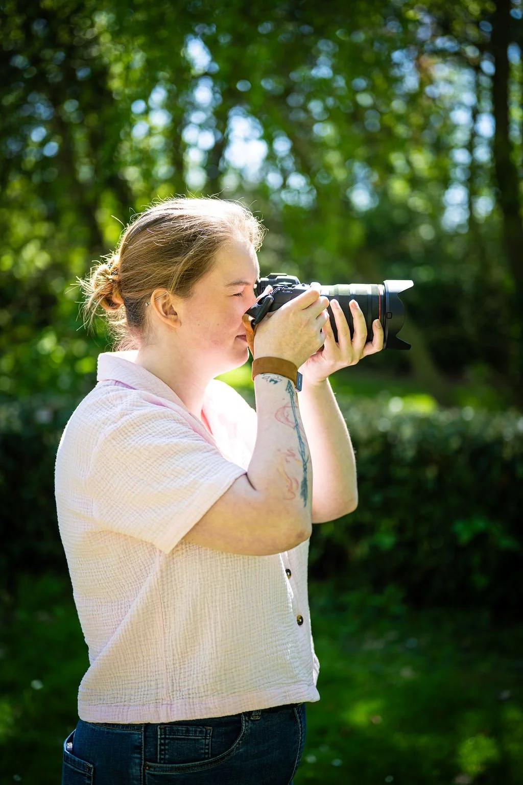 Vrouw met tatoeages op arm die een digitale camera vasthoudt en foto's maakt in een groene, zonnige buitenomgeving.