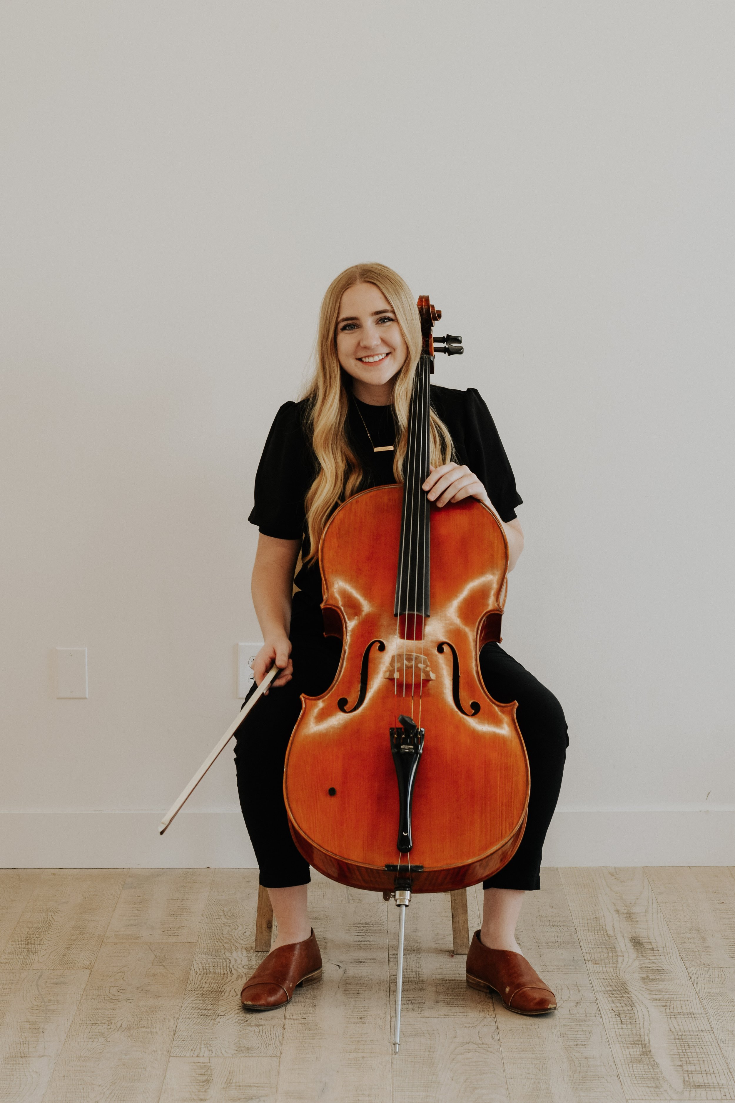 A woman with long blonde hair, wearing a black top and black pants, sitting on a wooden stool, holding a cello and bow, smiling, against a plain white wall.