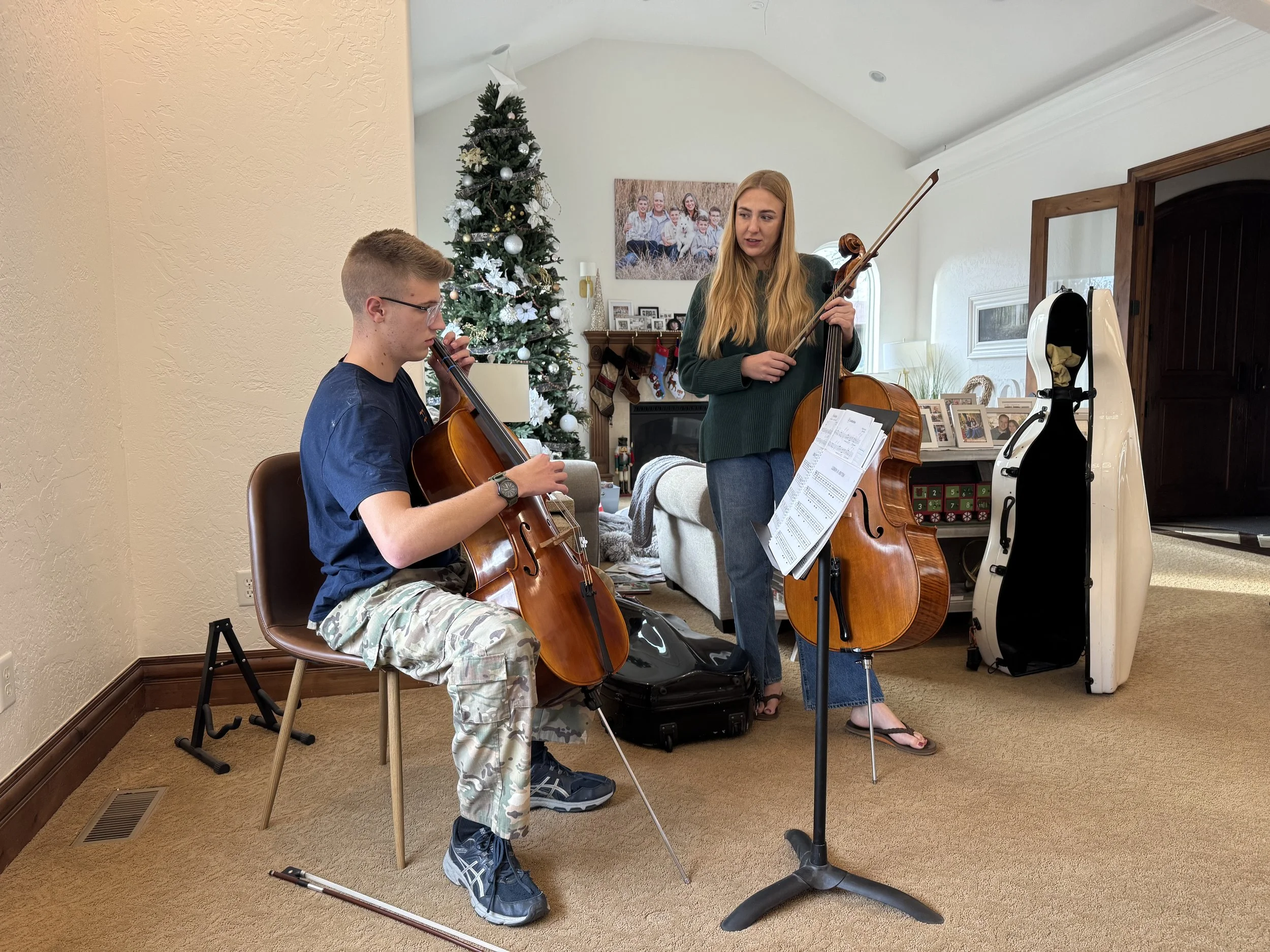 Two young musicians, a man and a woman, playing cellos indoors during Christmas time. The man is sitting and reading sheet music while playing, wearing glasses, a navy t-shirt, and camouflage pants. The woman is standing and explaining, holding her bow, wearing a dark green sweater and jeans. A decorated Christmas tree with ornaments and a star on top is in the background, along with family photos and Christmas stockings hanging on the fireplace.