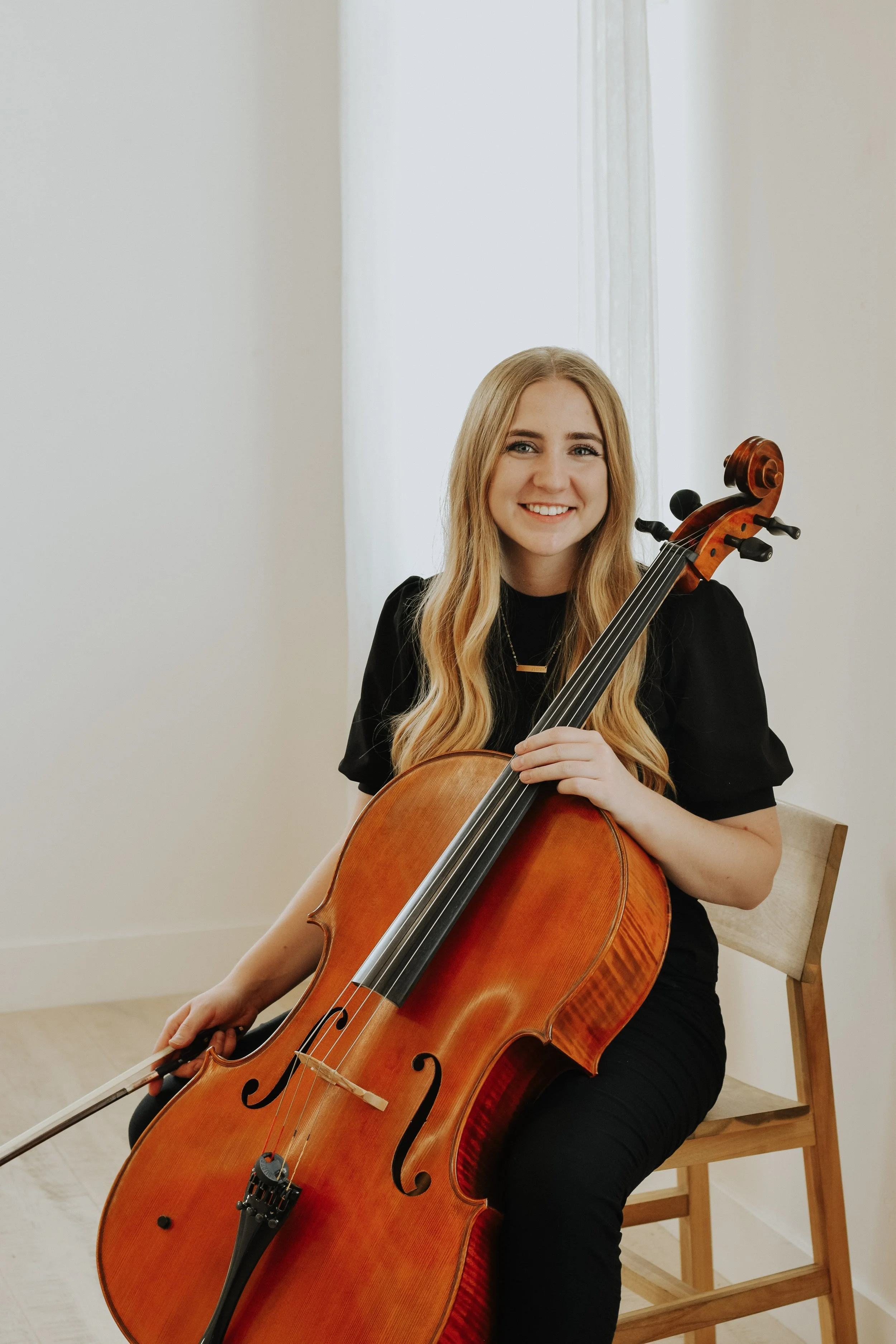 A young woman with long blonde hair and a black shirt sitting on a wooden chair, holding a cello, smiling at the camera, in a softly lit room with plain white walls and a light-colored floor.
