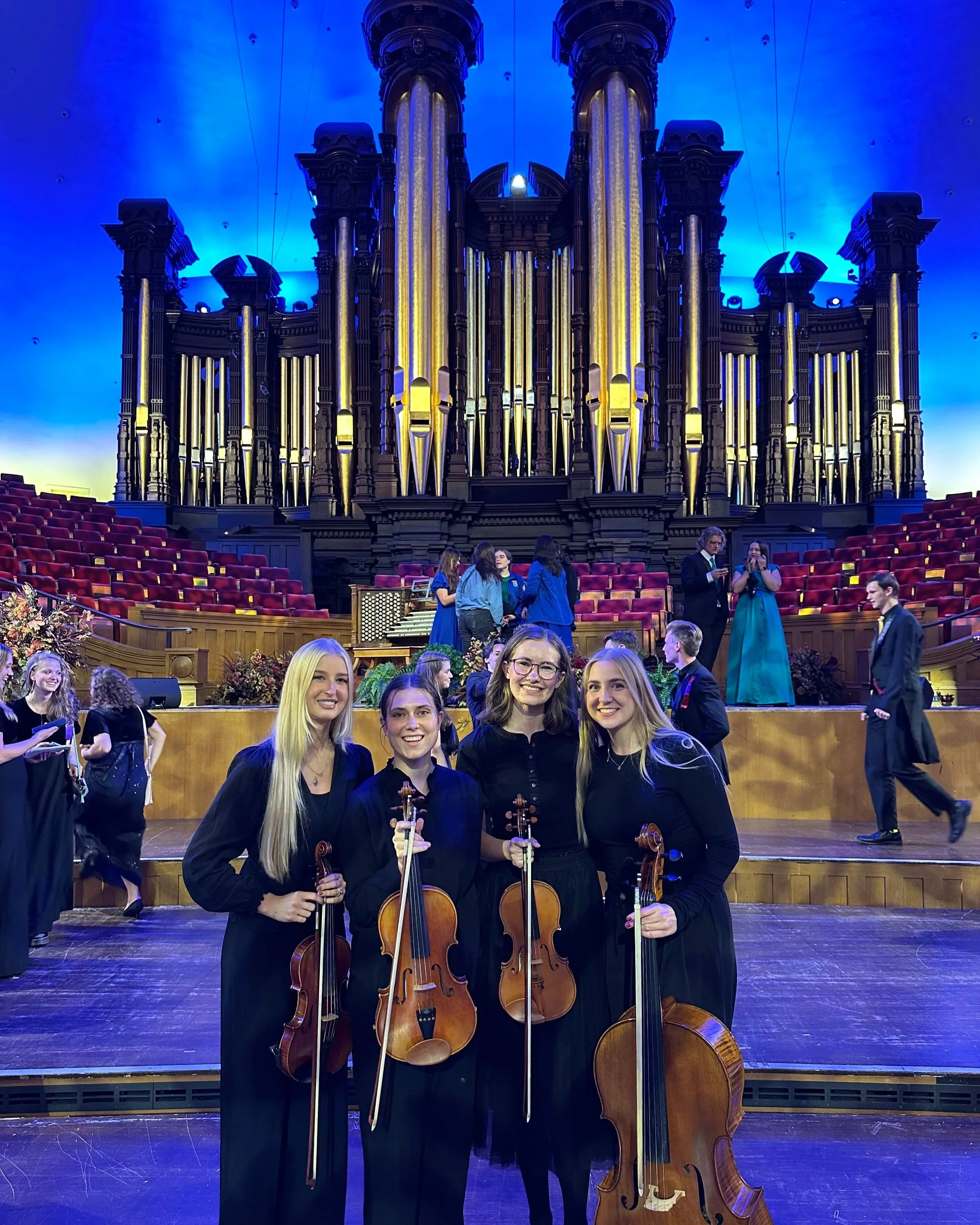 Young women holding violins and a cello on stage after a musical performance, in front of a large pipe organ with blue lighting.