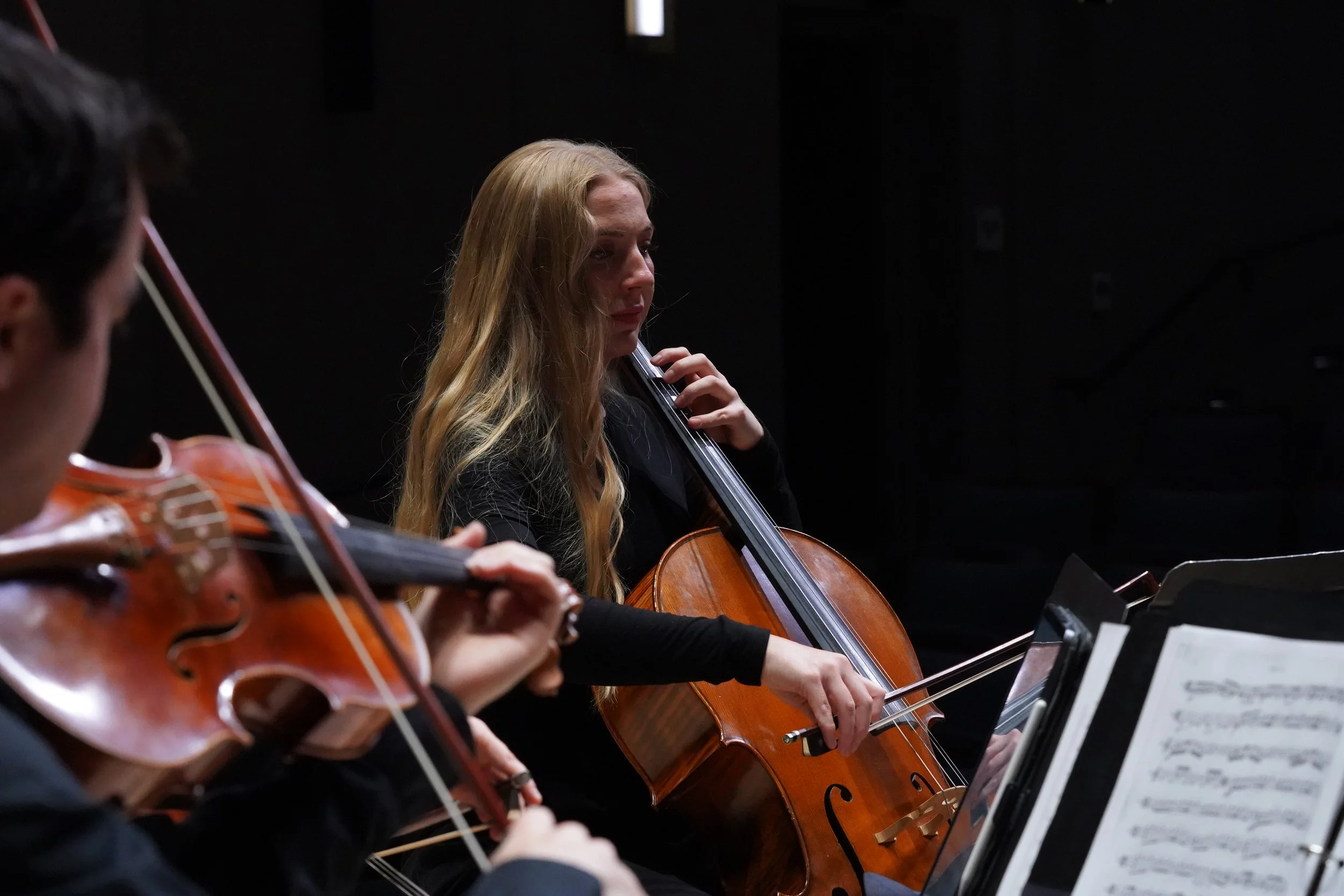 Two women playing cellos during a music rehearsal, with sheet music on stands in front of them in a dark room.