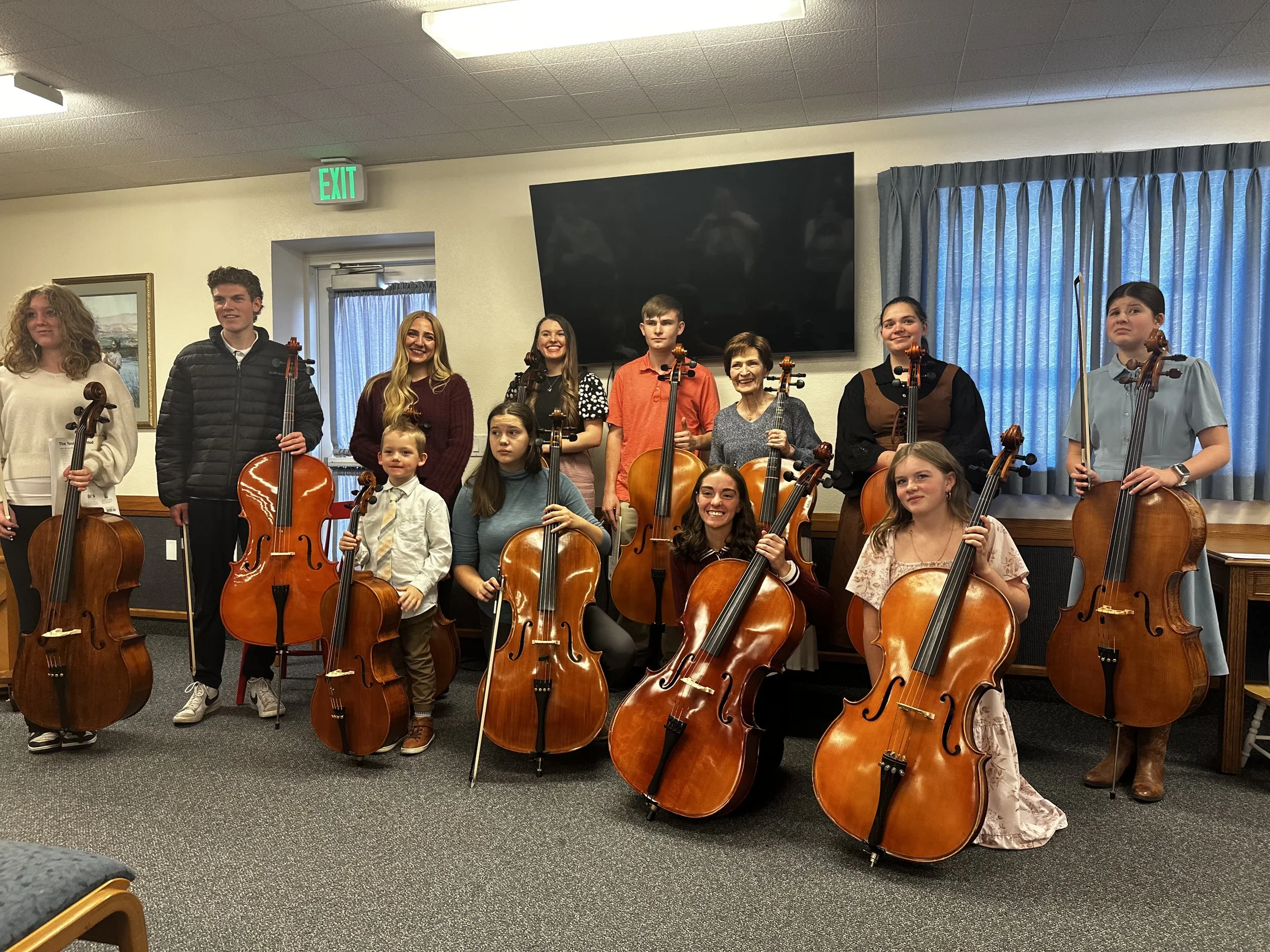 A group of children and young adults holding violins and cellos in a room with blue curtains and a large black television screen on the wall, smiling and posing for a photo.