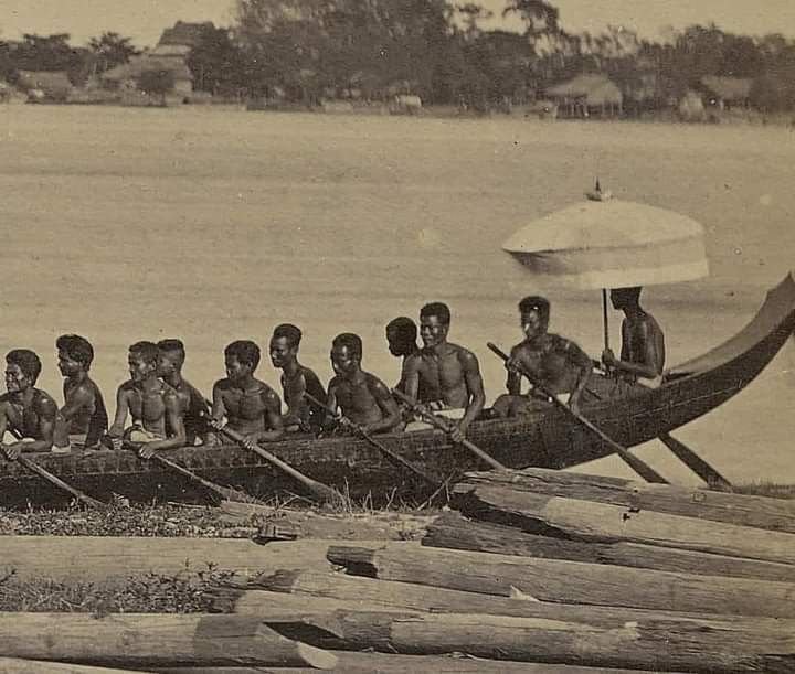 A group of young boys sitting in a traditional boat on a river, holding oars, with a person on the boat holding an umbrella. Wooden logs are in the foreground, and there is a small village or settlement on the opposite riverbank.