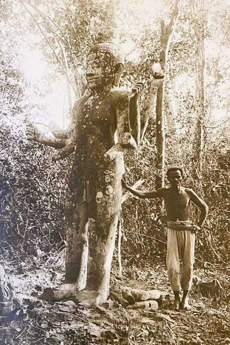 A large, ancient carved wooden statue of a humanoid figure with multiple arms, surrounded by dense forest. A shirtless man with striped pants stands beside the statue, touching its arm.