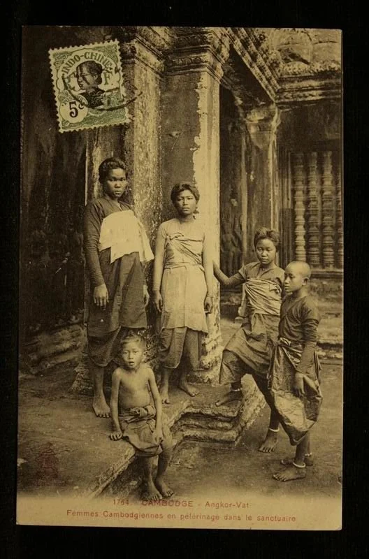 An old black-and-white photograph of five Cambodian women and children standing outside a temple or sanctuary with stone columns and intricate carvings. A vintage postcard stamp is in the top left corner. The caption indicates it shows Cambodian wome