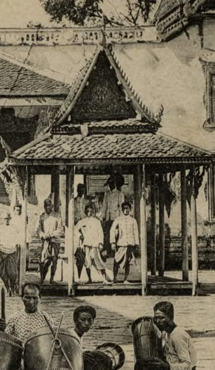 Historical black and white photograph of a group of children and a man standing and sitting outside a traditional building with a pointed roof and ornate decorations, possibly in a tropical setting.
