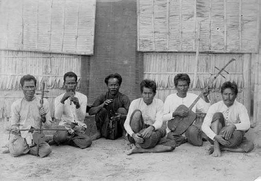 Six men sitting on the ground in front of a thatched wall, holding traditional musical instruments.