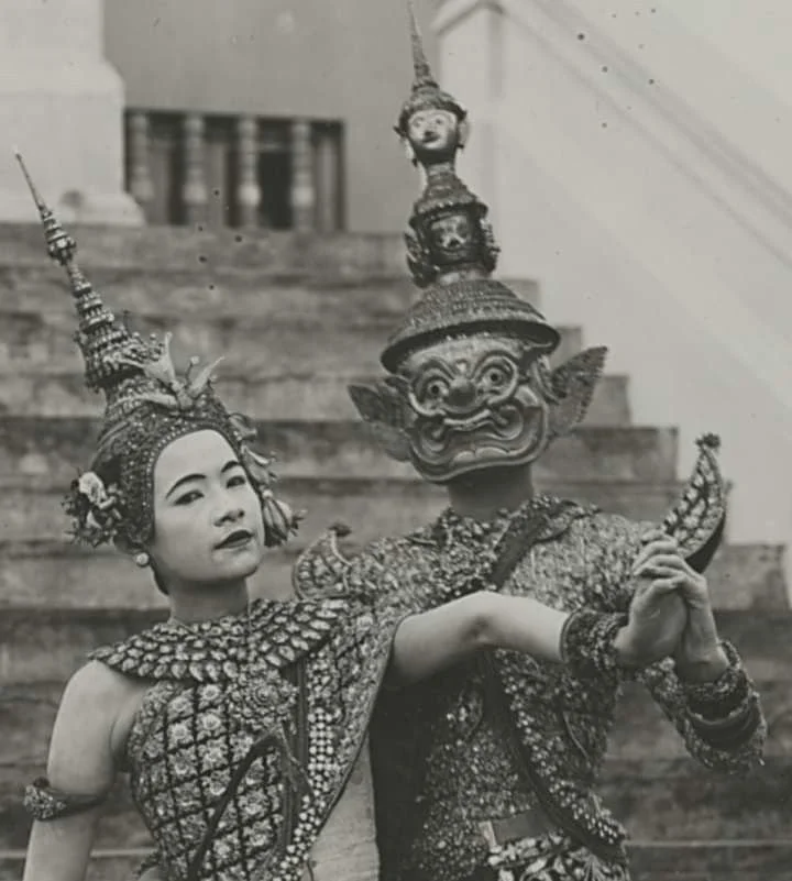A woman in traditional ornate costume and headdress poses with a person in a large puppet costume of a mythical creature, both standing on steps.