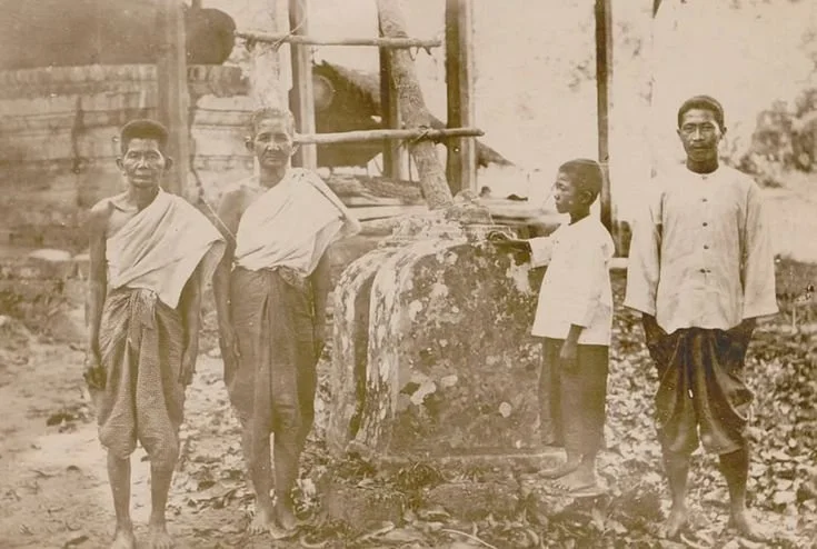 Four men are standing outdoors next to a structure with a stone base and wooden support beams. They are dressed in traditional clothing, with some wearing dhotis and shawls. The setting appears rustic with trees and a building in the background. The 