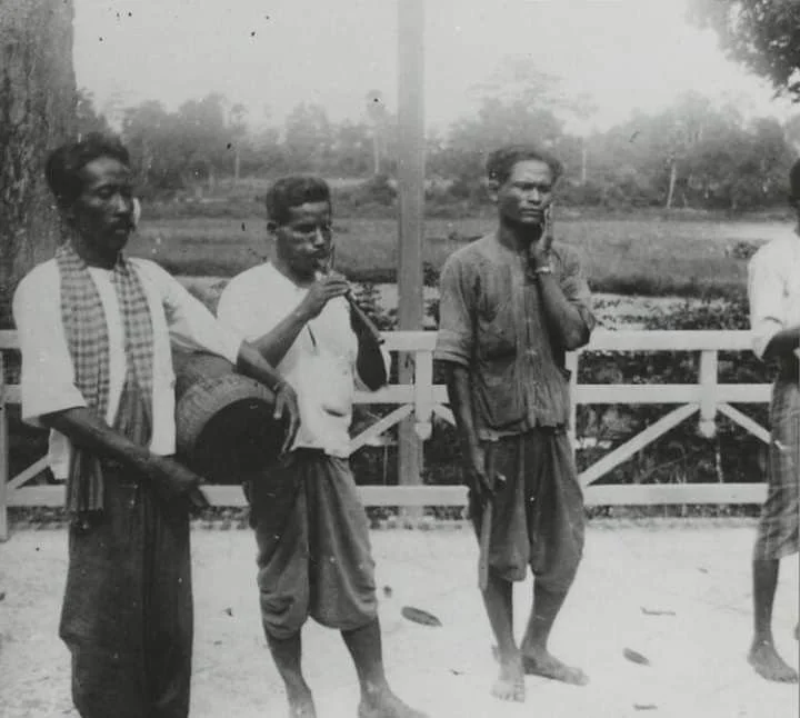 Four men standing outdoors near a wooden fence, with three of them preparing to perform, some holding musical instruments, in black and white.