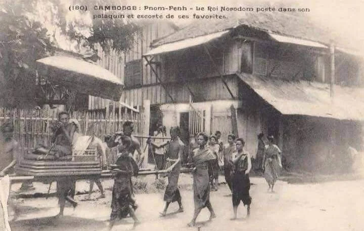Historical black and white photograph showing children and adults walking in front of a building with an awning. There are bamboo fences and a cart in the scene.