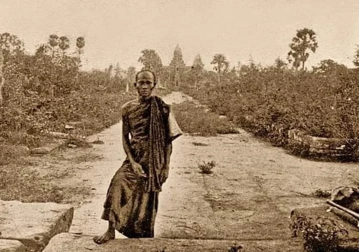 A young boy walking barefoot on a dirt path in a rural landscape with trees and a distant building in the background.