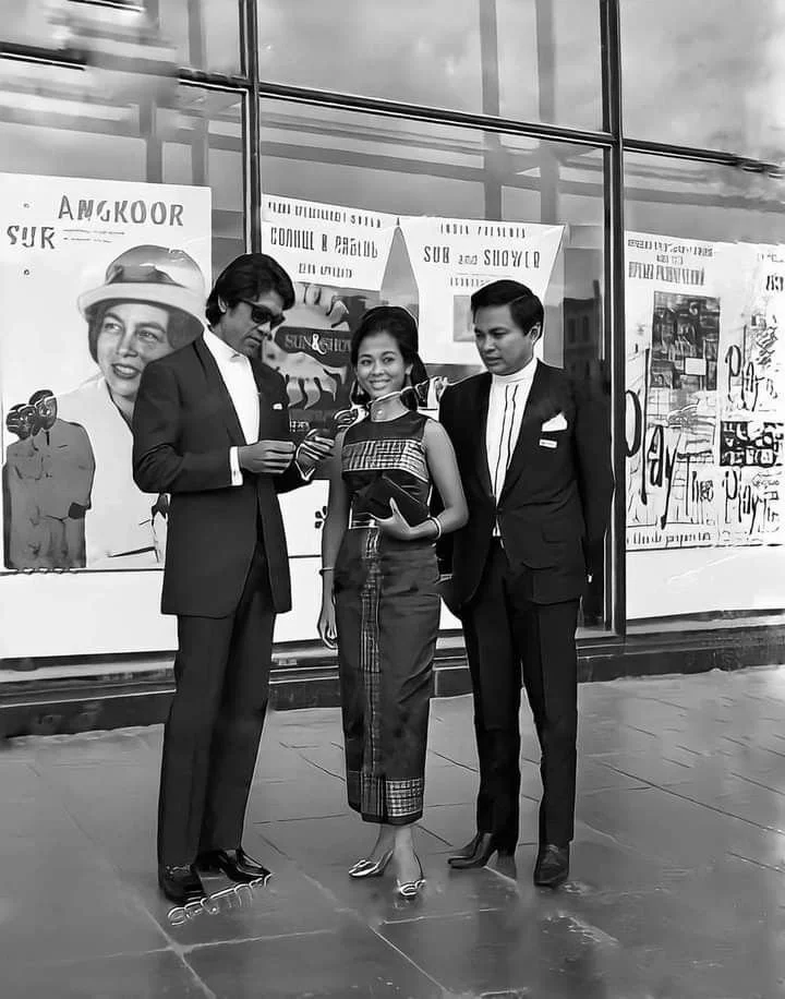Three people standing outside a storefront window, two men in suits and a woman in casual attire. The man on the left is holding a phone, the woman is smiling and holding a large clutch, and the man on the right is looking at the woman's phone.