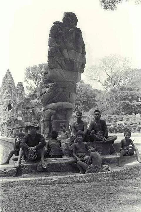 Group of children sitting and standing around a large stone sculpture of a standing figure at an outdoor historical site, with trees and ruins in the background.