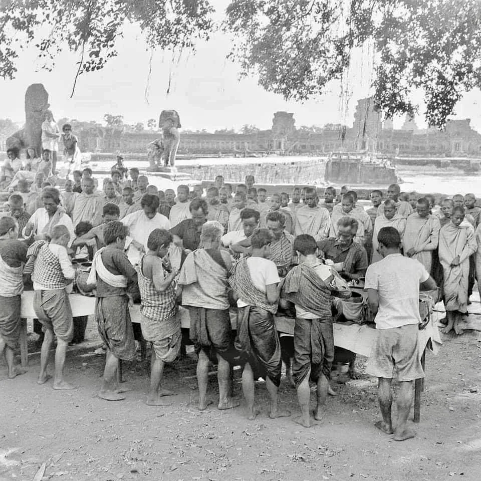 A group of children and adults gathered outdoors around a table, likely distributing or receiving food, with a historical site and ruins in the background.