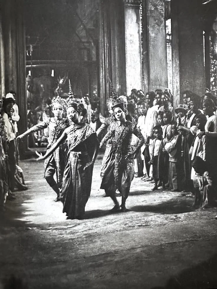 Gallery scene with three women in traditional costumes performing a dance in front of a large crowd, black and white photograph.