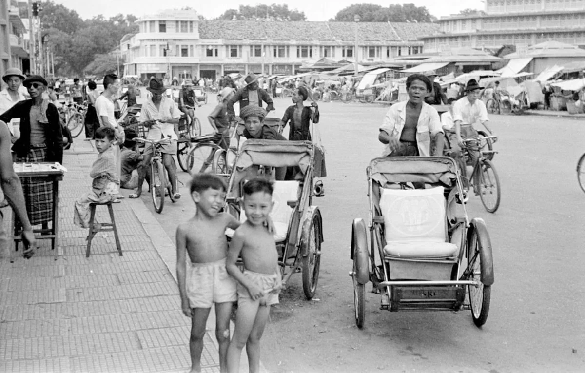 Black and white photograph of a busy urban street scene with children and adults, rickshaws, bicycles, and market stalls in the background.