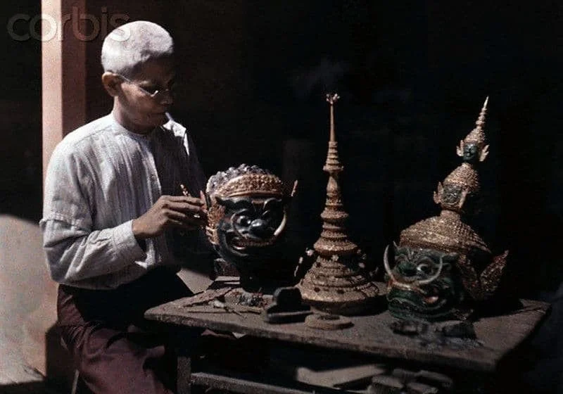 A person wearing glasses and traditional clothing sits at a table with ornate, carved masks and statues, including two large face masks and a tiered, decorative structure, in a dimly lit environment.