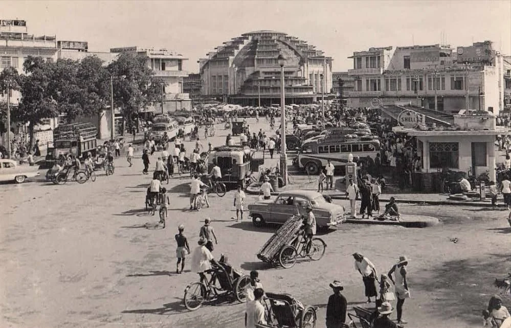 Black and white photo of a busy marketplace with cars, bicycles, pedicabs, and people on the street, with a large modern building in the background.