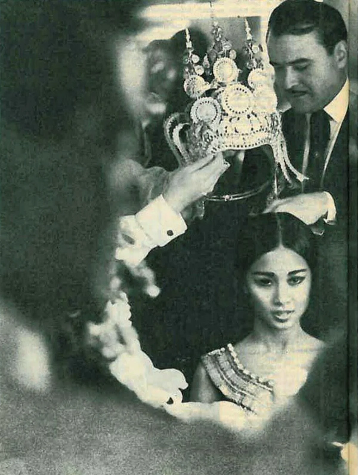 Black-and-white photograph of a woman in a headdress being presented to a man in a suit by other people, with a woman in the foreground.