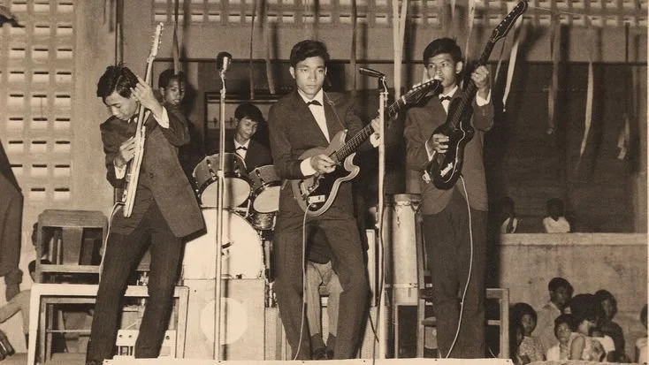 Four young male musicians performing on stage with guitars and drums, dressed in formal suits, in front of an audience.