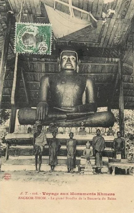 Black and white photo of five young boys standing in front of a large seated Buddha statue under a thatched roof, with a boat hanging from the ceiling.