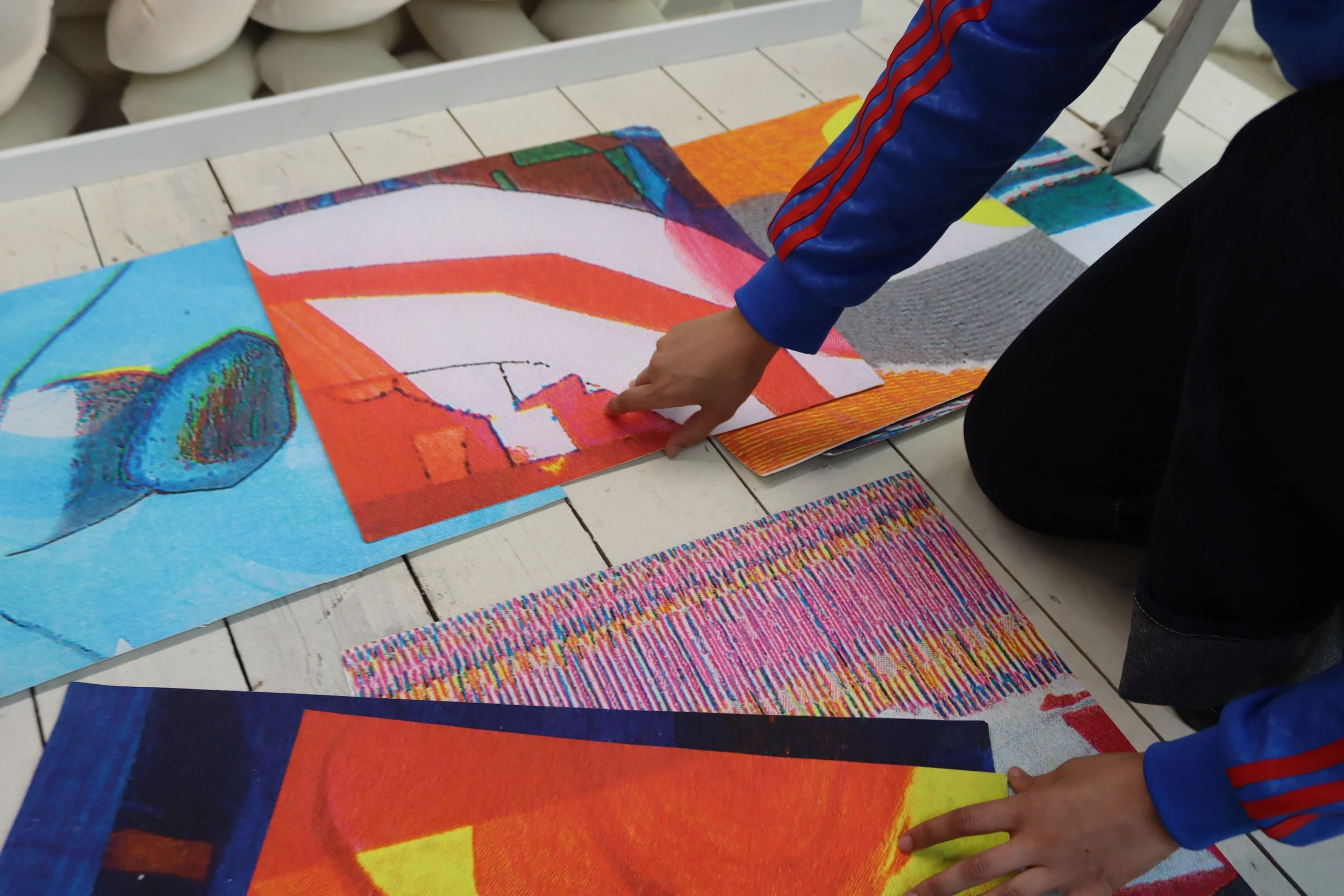 Artist in a blue jacket with red stripes is arranging colorful abstract art prints on a white wooden floor.