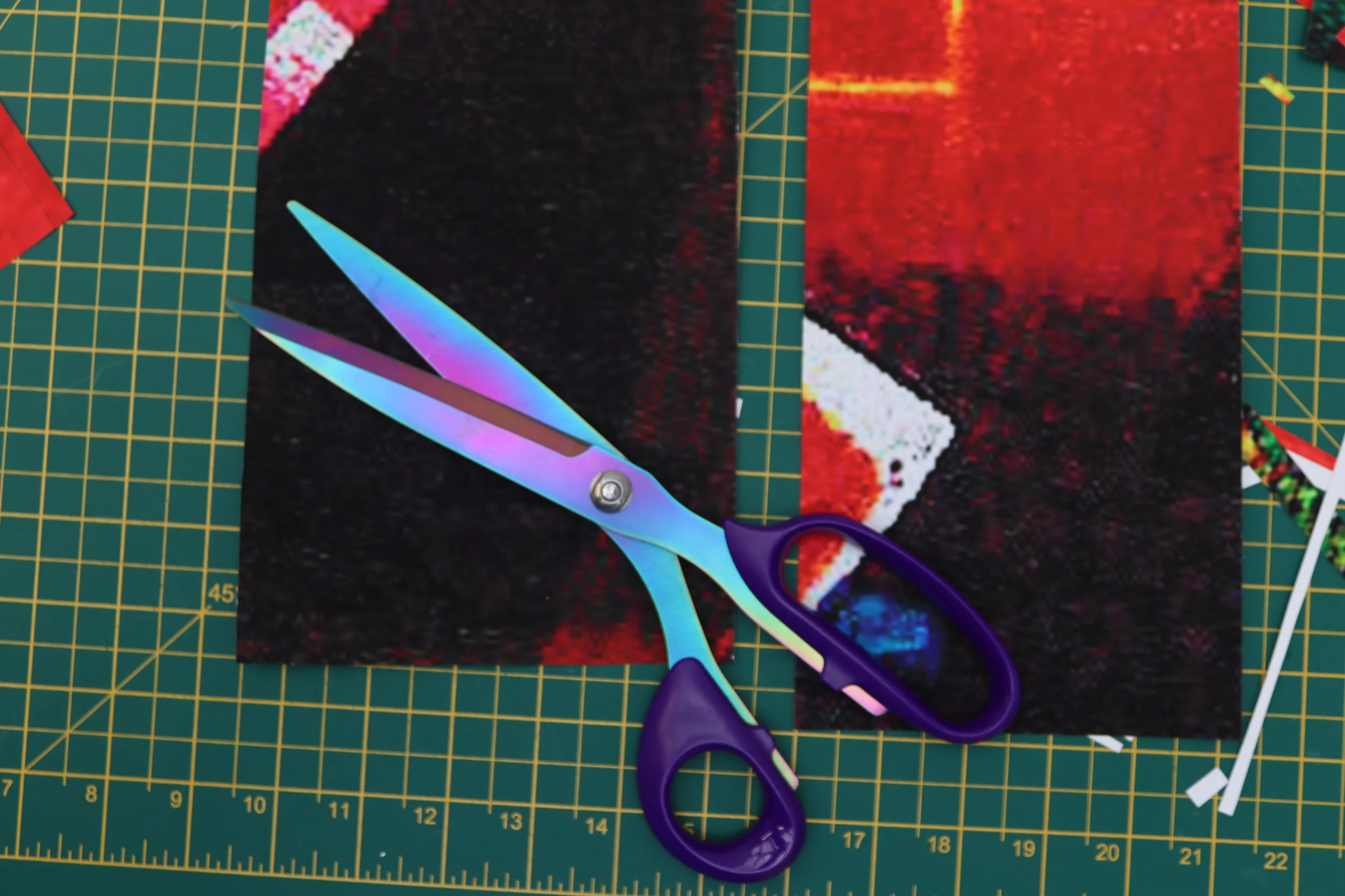A pair of iridescent scissors with purple handles resting on a cut print with a red, black, and white geometric pattern, surrounded by miscellaneous paper scraps and a green cutting mat with yellow grid lines.