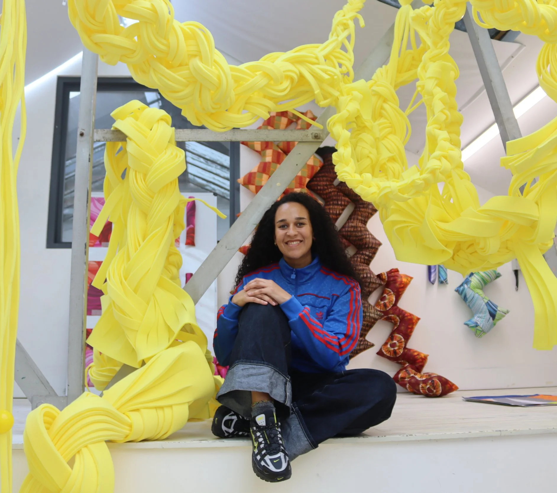 A woman sitting on the floor in front of a vibrant display of yellow, colourful installation art, smiling at the camera.