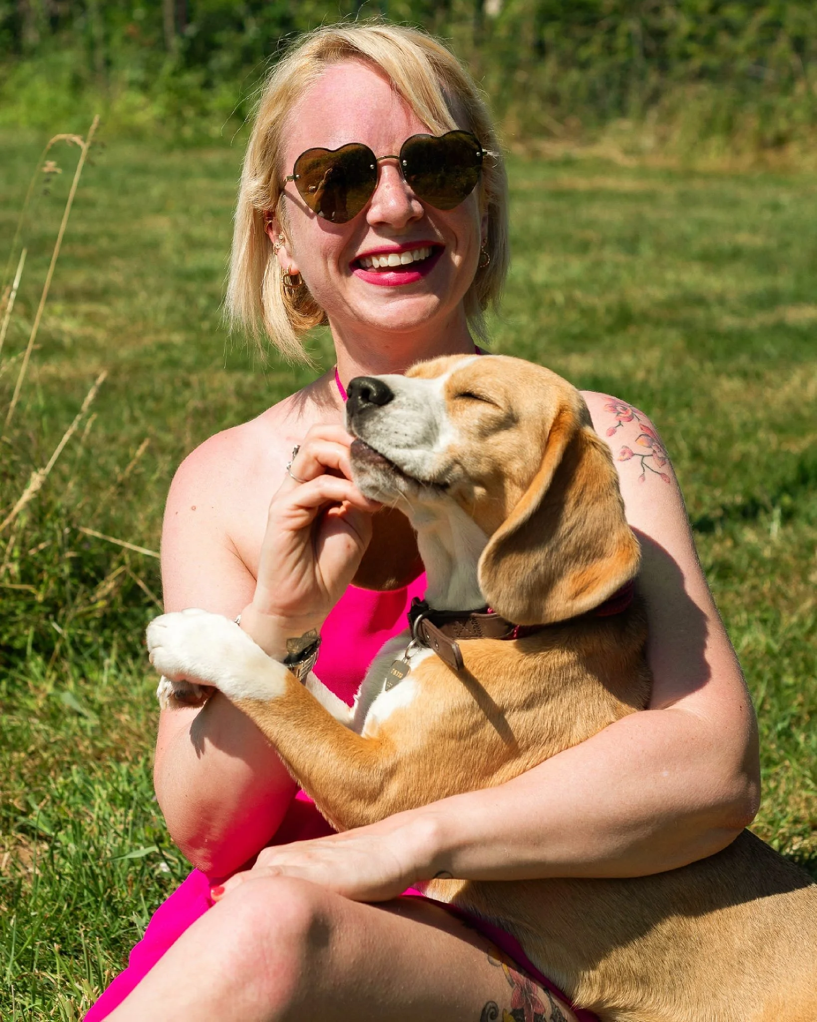 Une femme blonde portant des lunettes de soleil en forme de cœur, souriante et tenant un chien de race beagle, dans un champ verdoyant lors d'une journée ensoleillée.