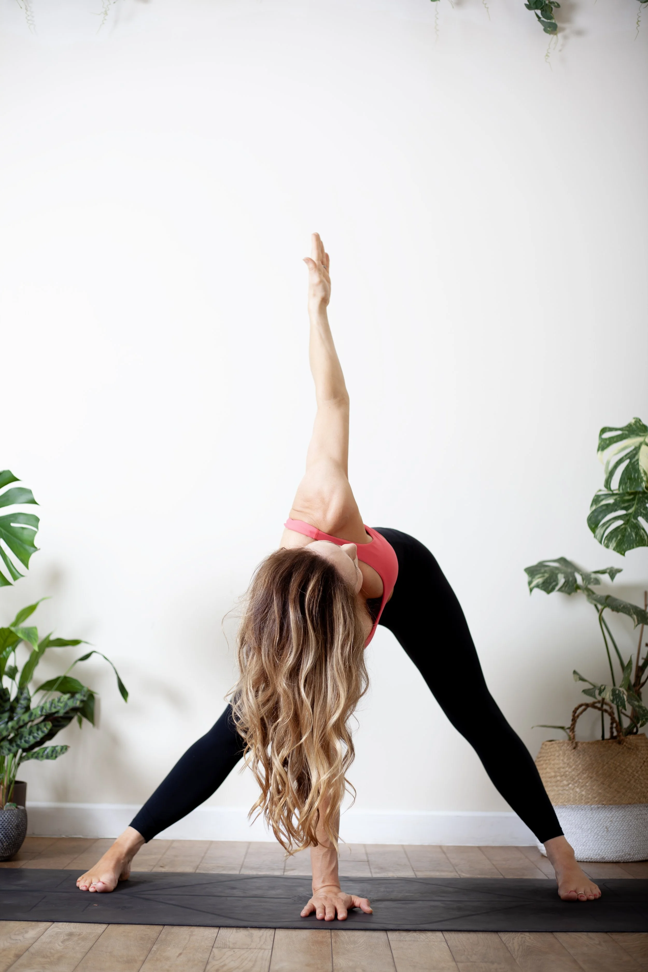 A woman with long, wavy blonde hair practicing yoga in a lunge pose inside a bright room with potted plants on either side.