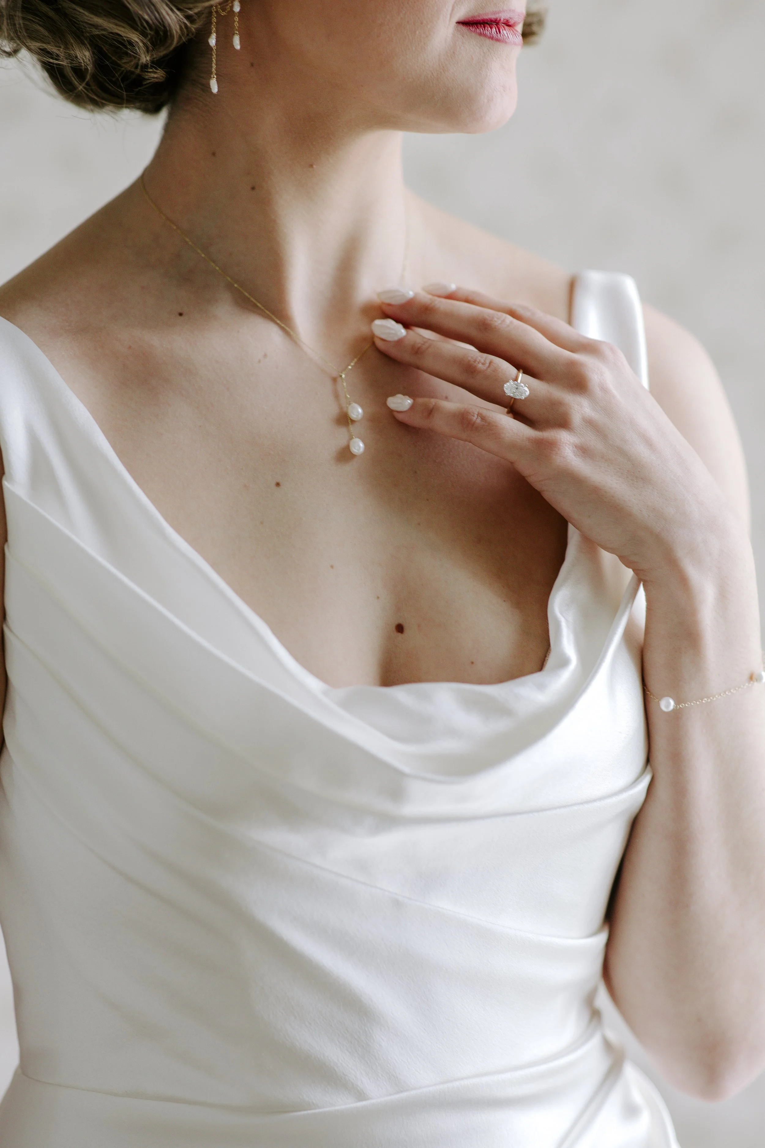 Close-up of a woman wearing a white satin dress with a V-neckline, adorned with pearl jewelry including a necklace, earrings, and rings, with her hand placed on her chest.