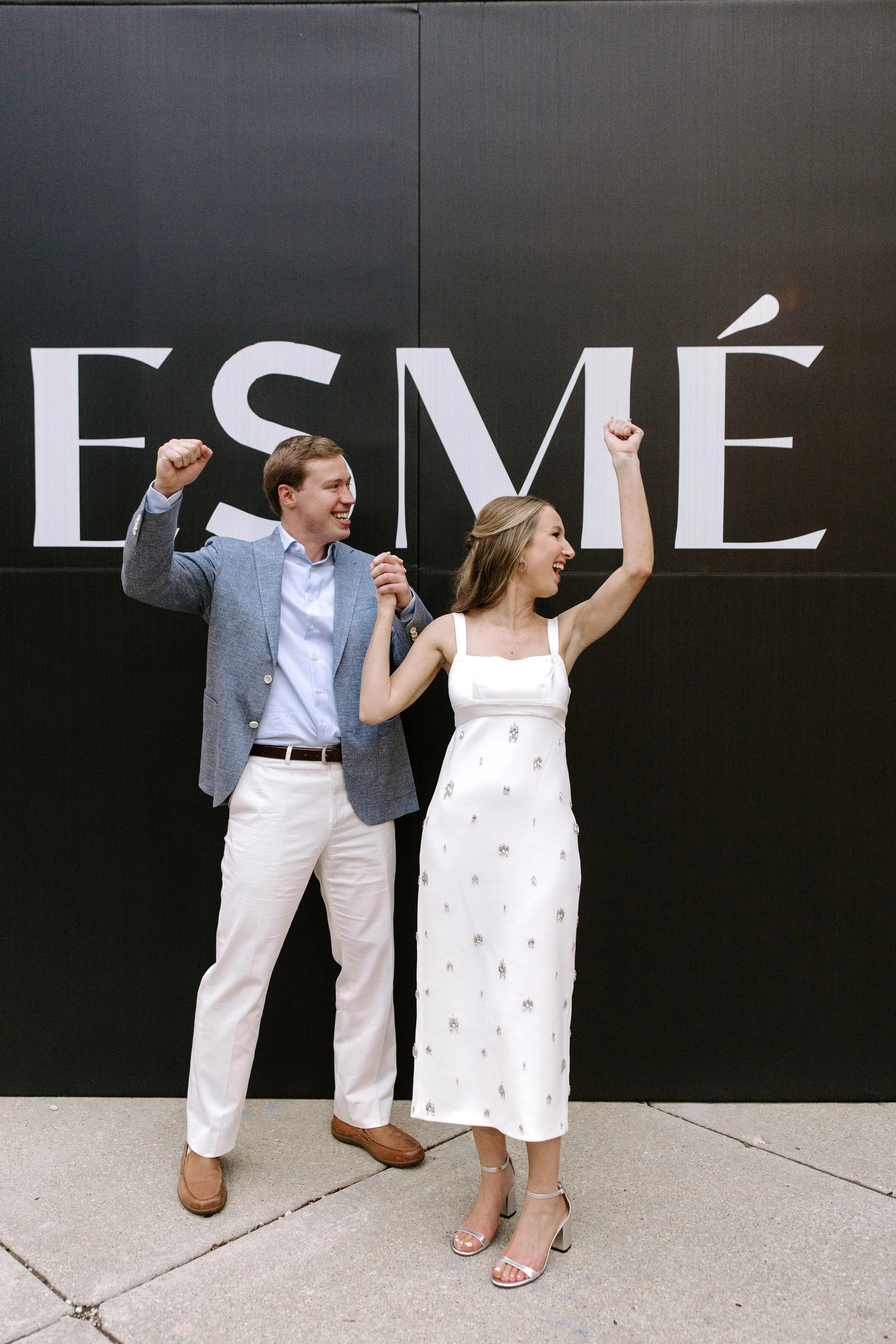 A man and woman celebrating in front of a black wall with large white text that reads 'ÉSMÉ'. The man is dressed in a gray blazer, white shirt, white pants, and brown shoes, with his fist raised and smiling. The woman is wearing a white dress with si