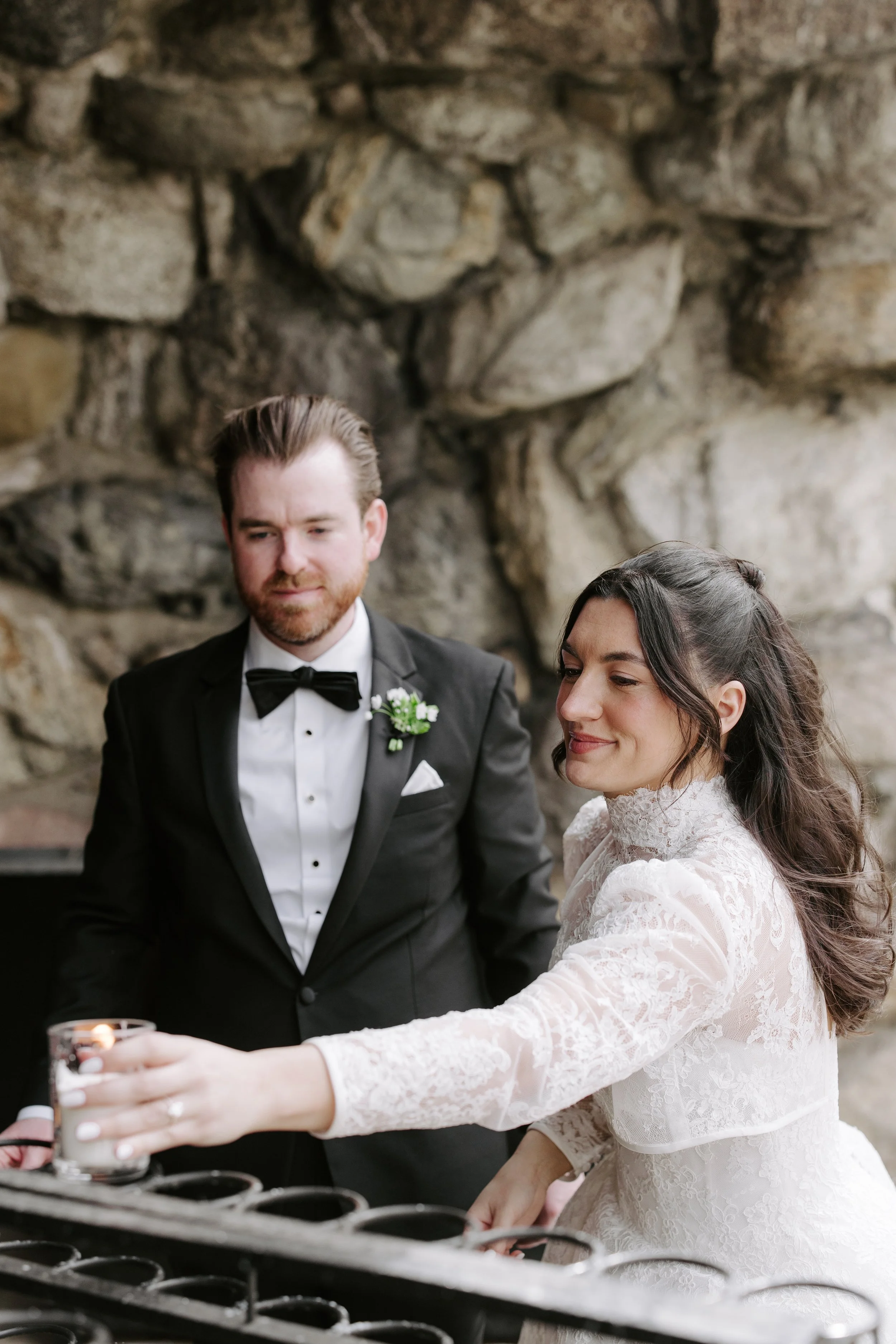 A newlywed couple in wedding attire participating in a candle lighting ceremony, with a stone wall in the background.