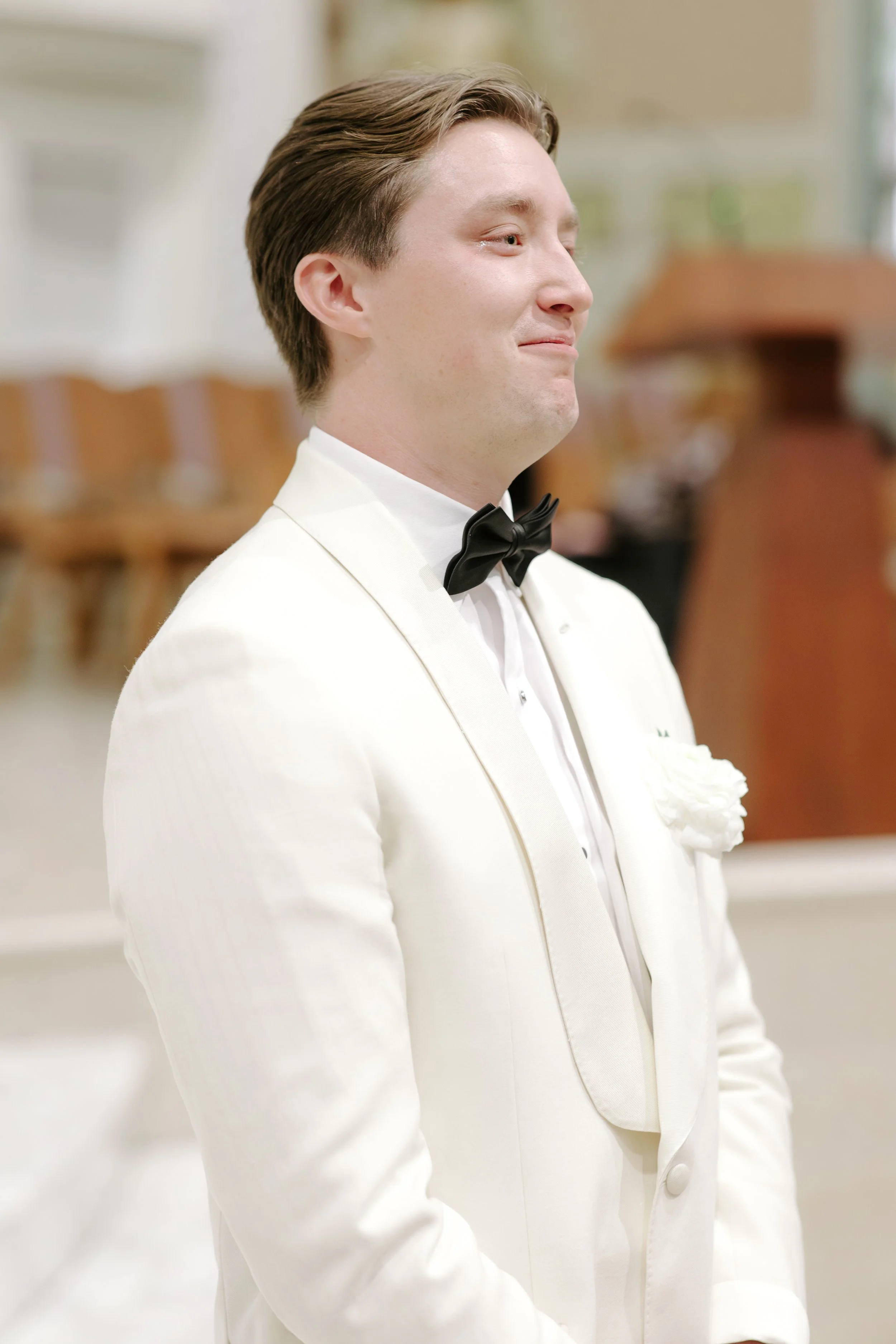 A man in a white tuxedo with a black bow tie and a white flower boutonniere, smiling softly, at a formal event.