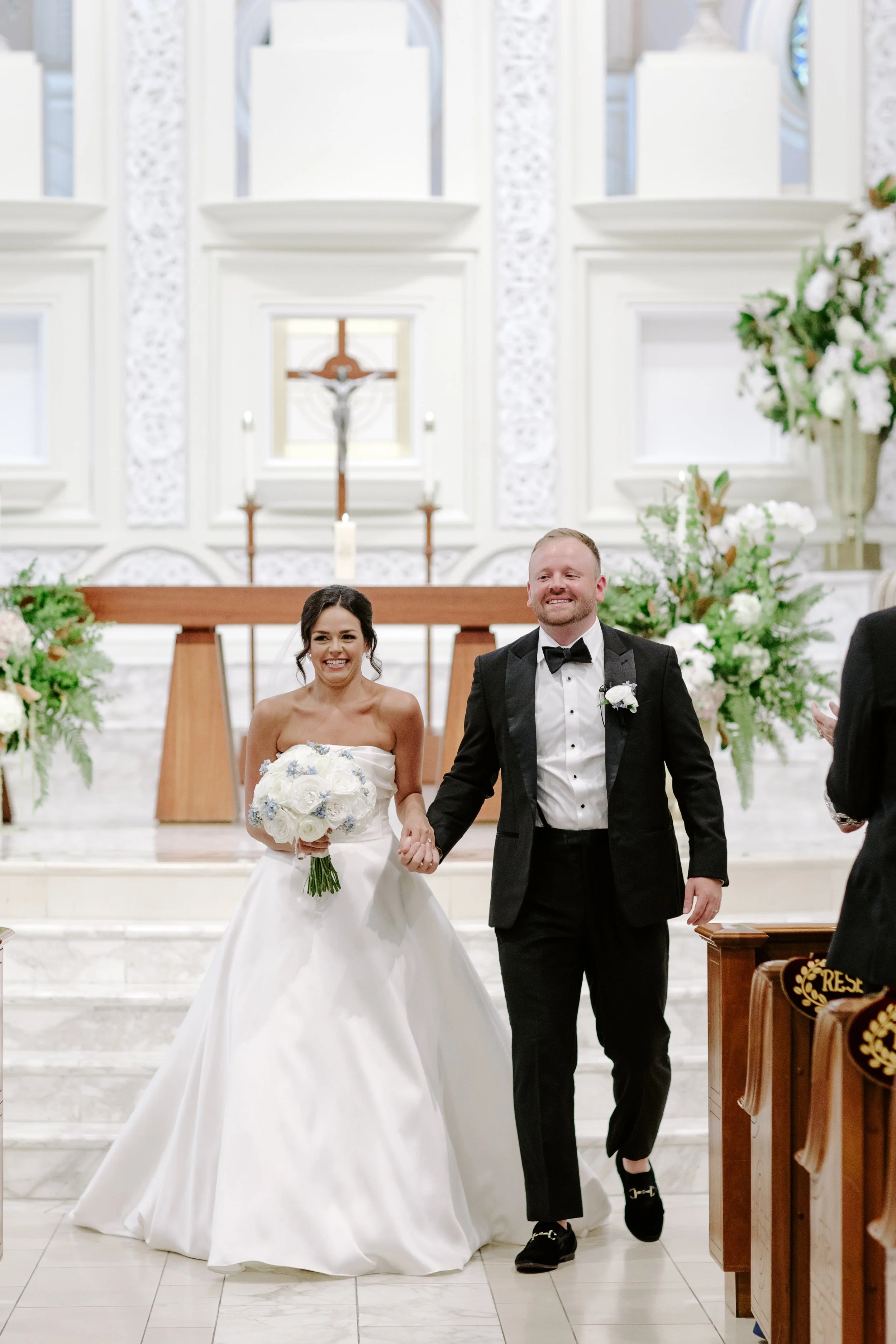 A newlywed couple holding hands and smiling in a church during their wedding ceremony. The bride is in a white wedding dress holding a bouquet of white flowers, and the groom is in a black tuxedo with a bow tie. The church features white walls, flora