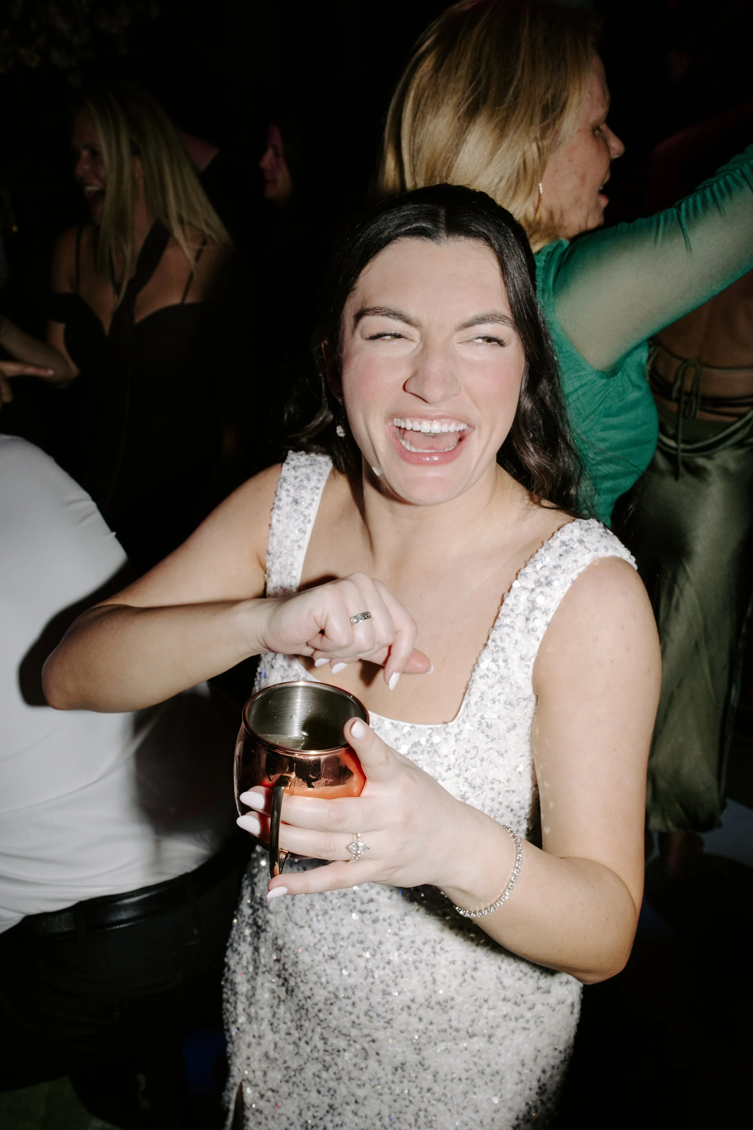 A woman in a white sequin dress holding a copper mug, smiling and laughing at a lively party.