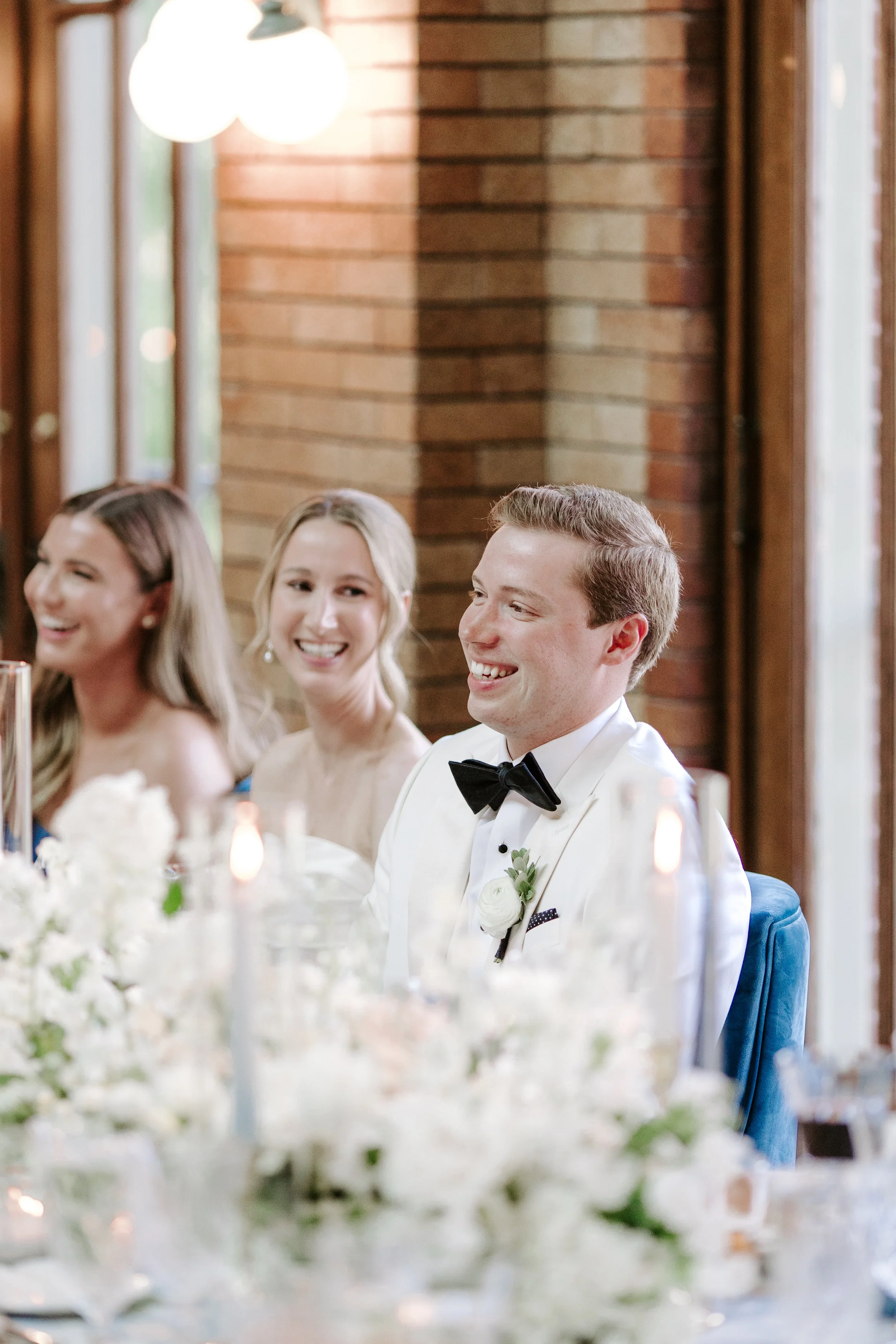 Smiling man in a tuxedo with a boutonniere at a wedding reception, seated at a table adorned with white flowers.