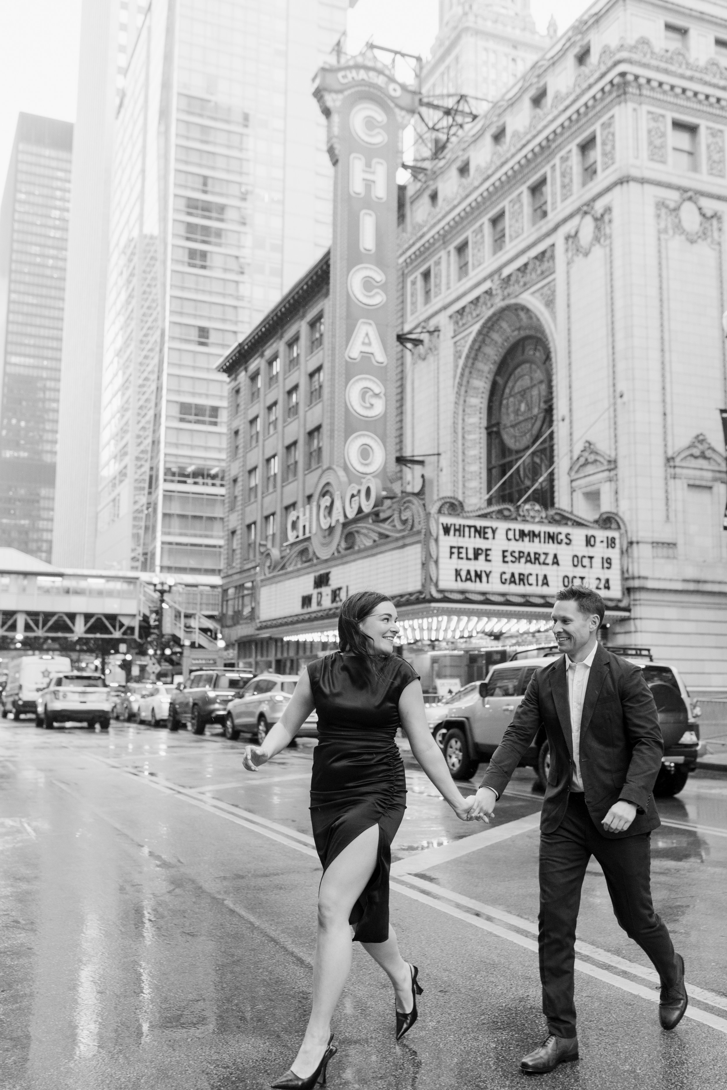 A man and woman holding hands and walking across a city street in front of a theater marquee and skyscrapers. The woman wears a black dress with high heels, and the man is in a suit. The theater sign reads 'CHICAGO' and the marquee shows upcoming per