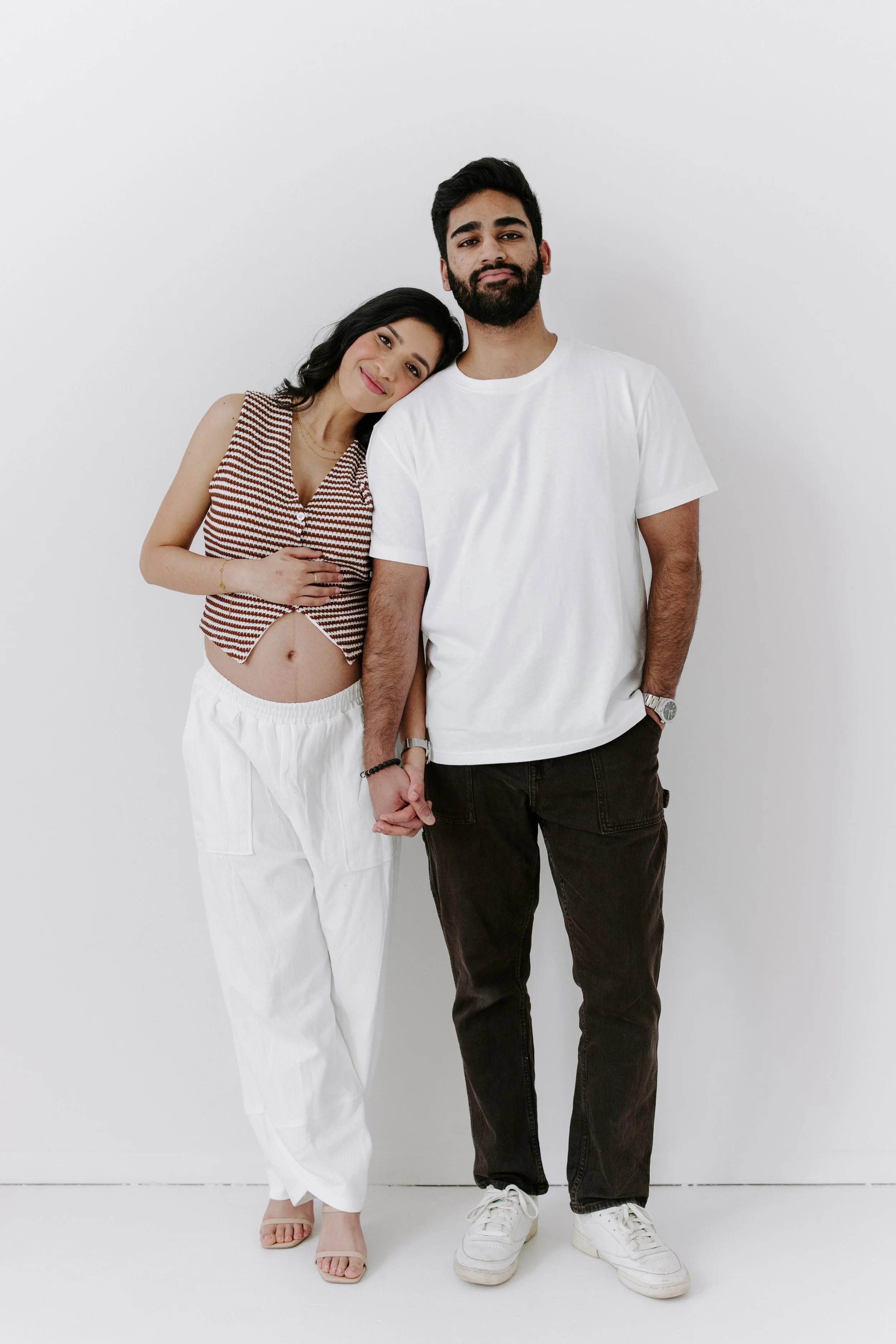 A woman with long dark hair wearing a sleeveless striped top and white pants holding hands with a bearded man wearing a white t-shirt and black pants, standing against a plain white background.