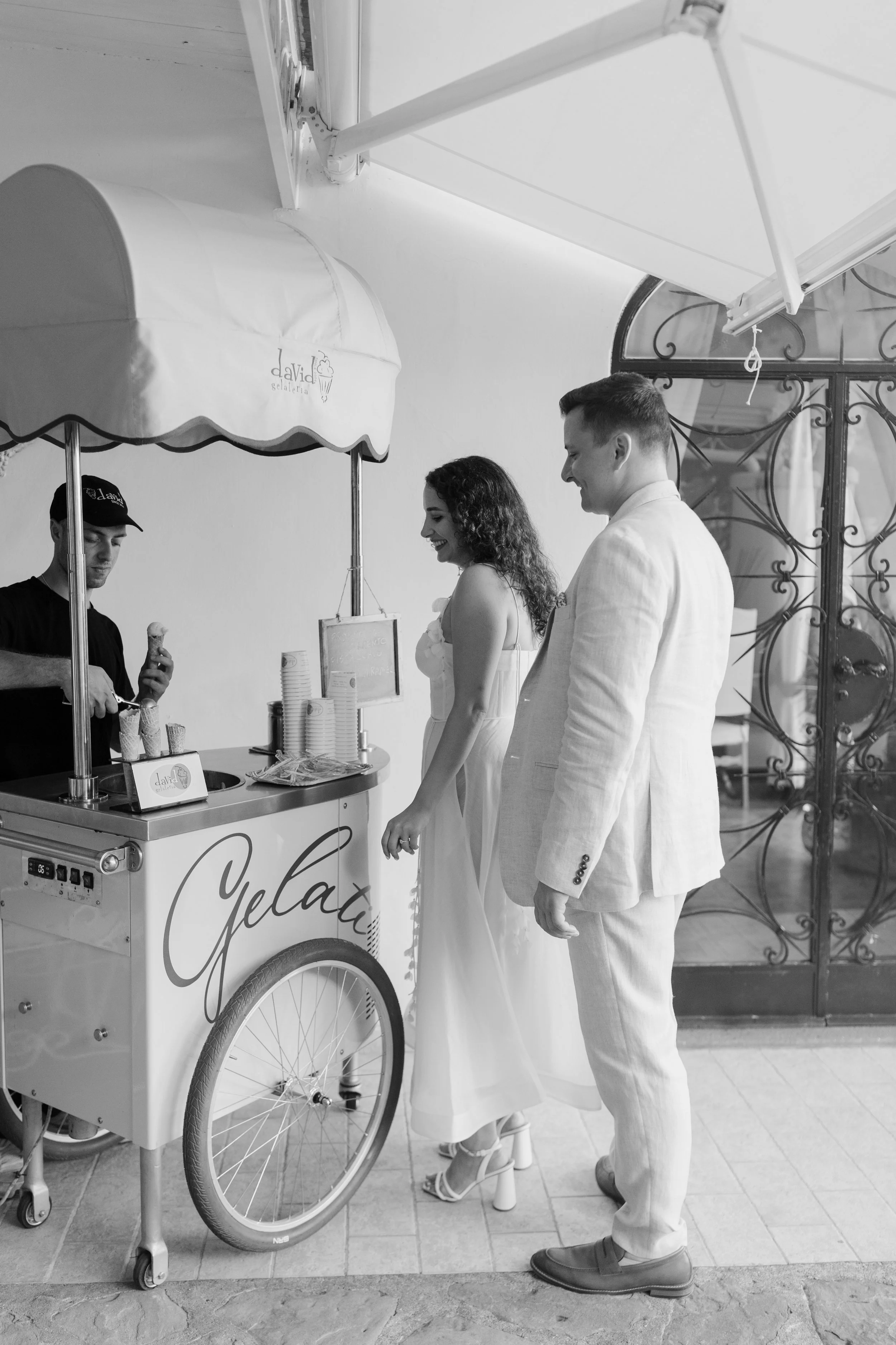 A woman and a man in light-colored suits standing next to an ice cream cart with a large umbrella, with a man in black serving ice cream from the cart.