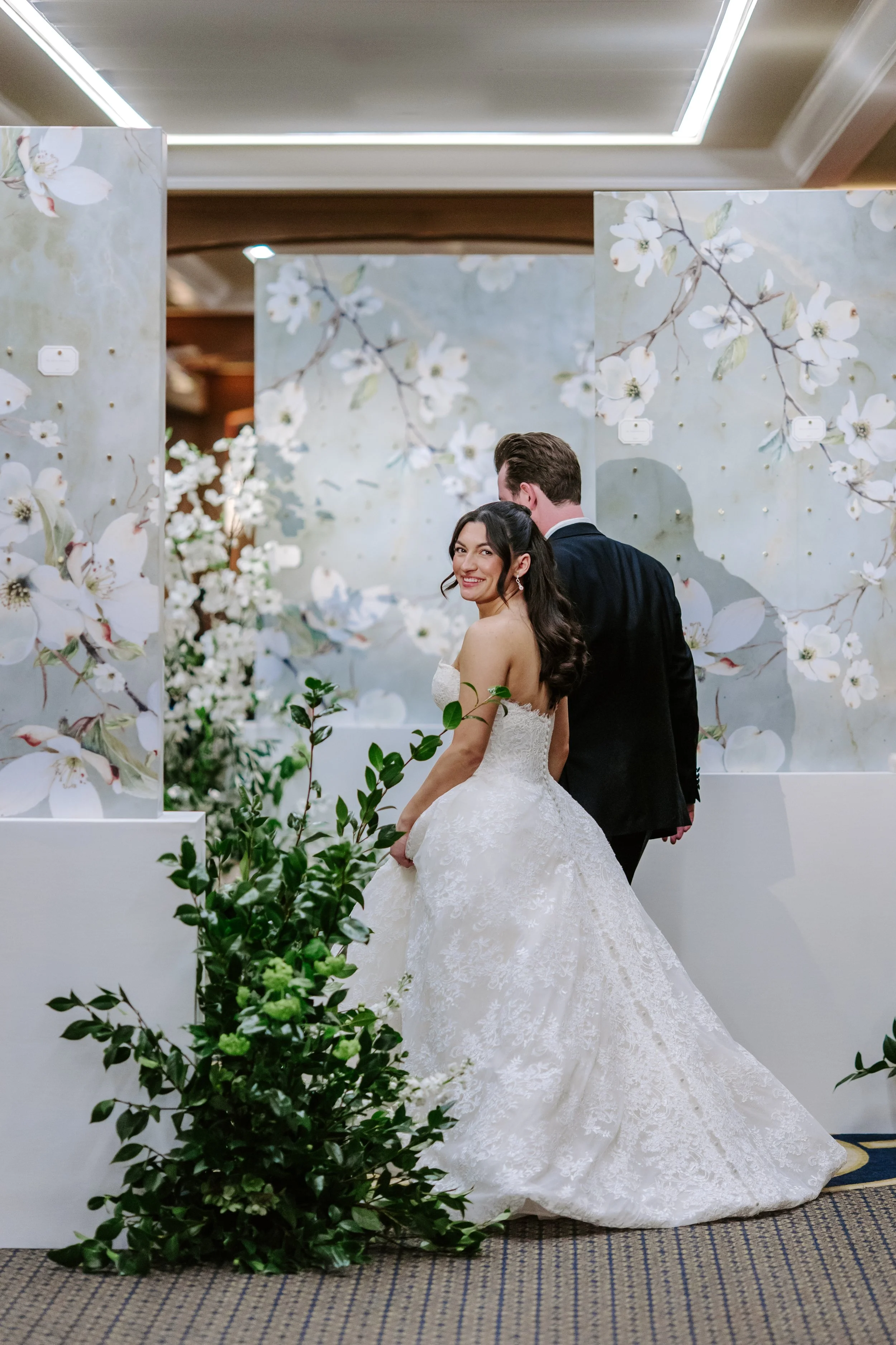 A bride in a white lace wedding gown smiling at the camera and standing next to a groom in a black suit. They are indoors with a decorative backdrop of white flowers and branches, and greenery in the foreground.