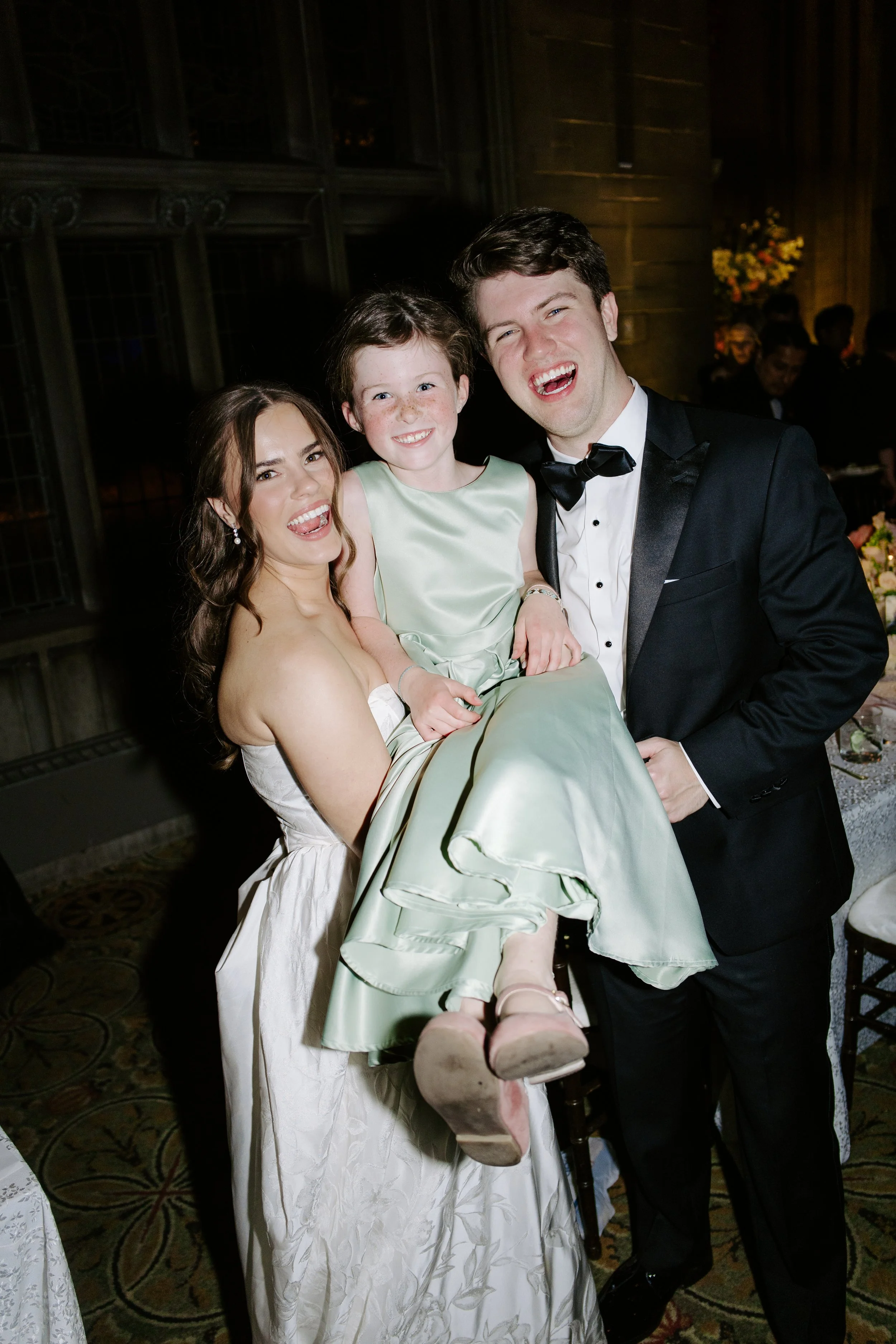 A woman in a white dress holding a young girl in a light green dress, and a man in a black tuxedo, all smiling and laughing at an indoor event with a dark wooden interior and floral arrangements in the background.