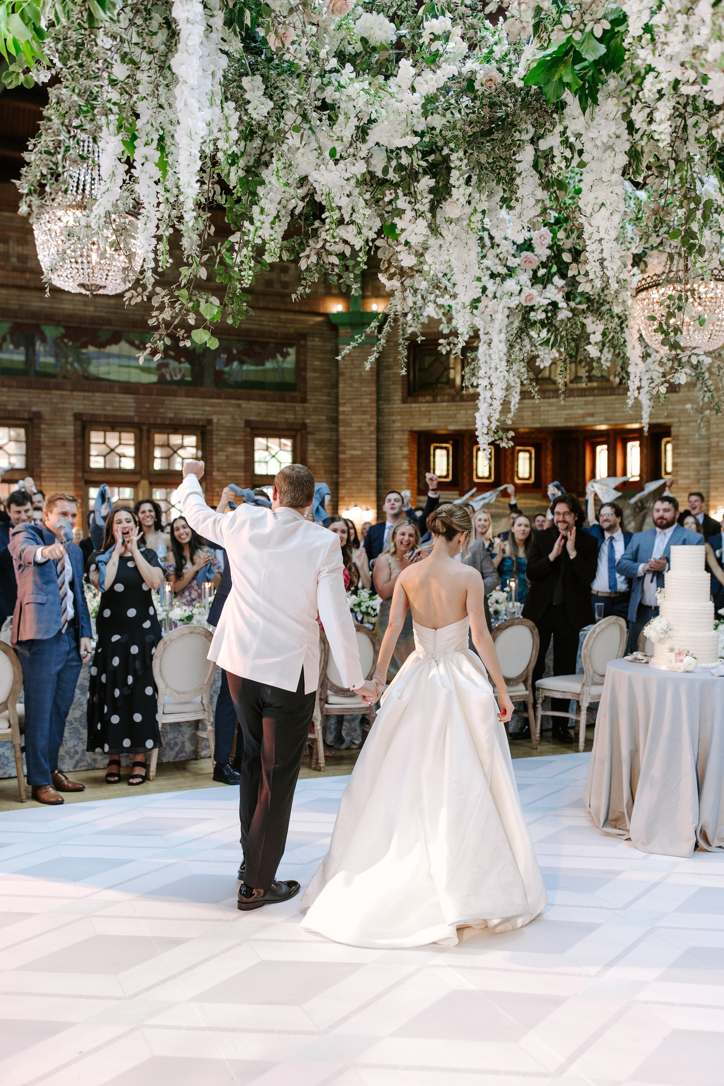 A newlywed couple is dancing at their wedding reception, surrounded by guests in a decorated venue with floral ceiling installation and chandeliers.