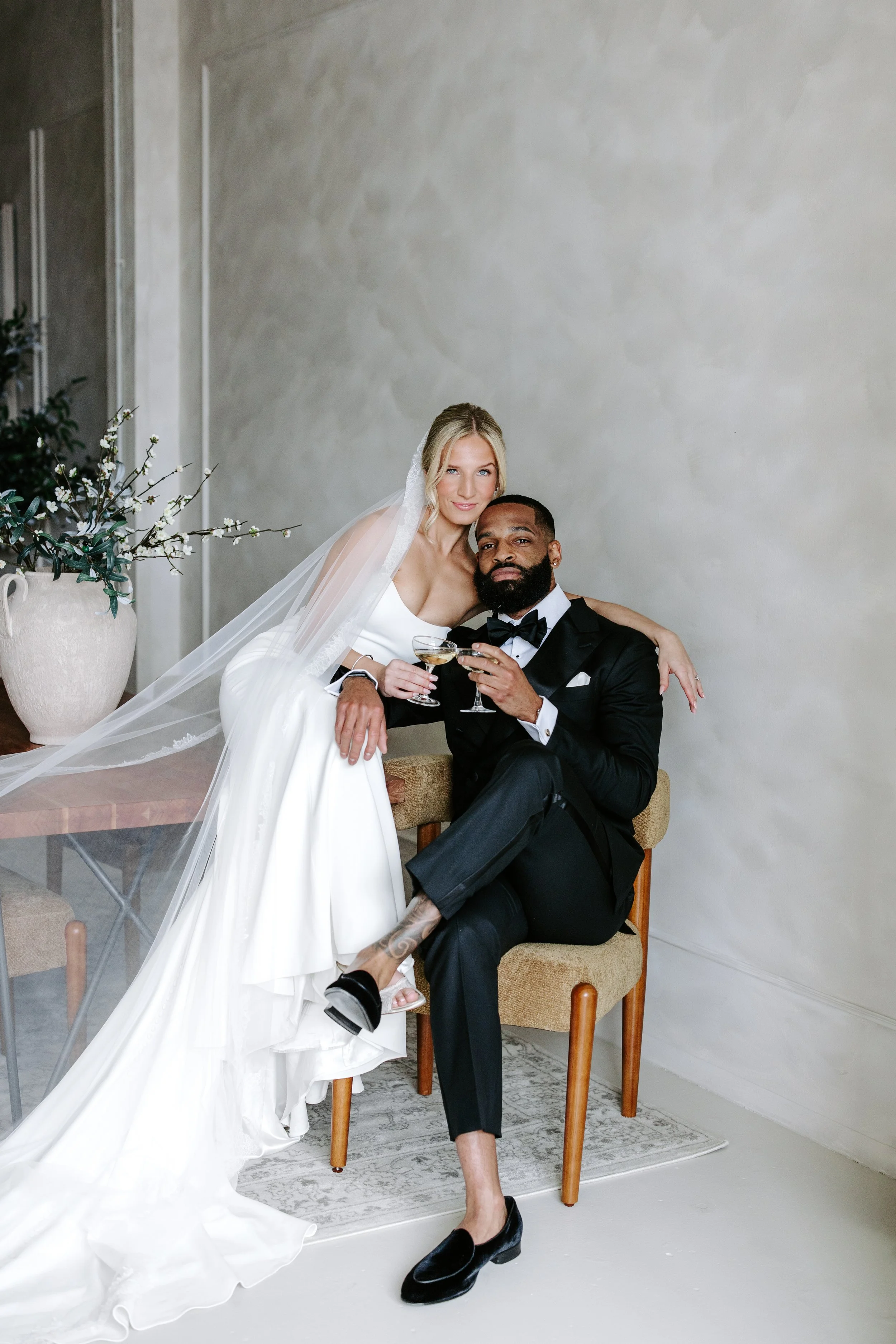 A bride in a white wedding dress and veil sitting on a chair with a groom in a black tuxedo and bowtie, holding a champagne glass, in a room with light-colored walls and a large floral arrangement.